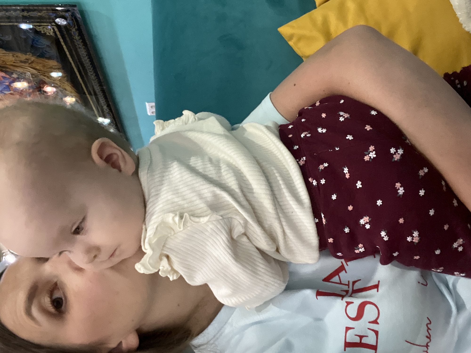The image shows a close-up of a woman holding a baby. The woman is kissing the baby on the cheek. The baby is wearing a cream-colored ribbed top with ruffled sleeves and maroon pants with a floral pattern. The woman is wearing a light blue t-shirt with red text on it. In the background, there is a turquoise couch and a yellow pillow. A framed artwork with lights is visible above the couch. The overall scene appears to be indoors. This image does not contain information that allows for the determination of its location in Moscow, Russia.