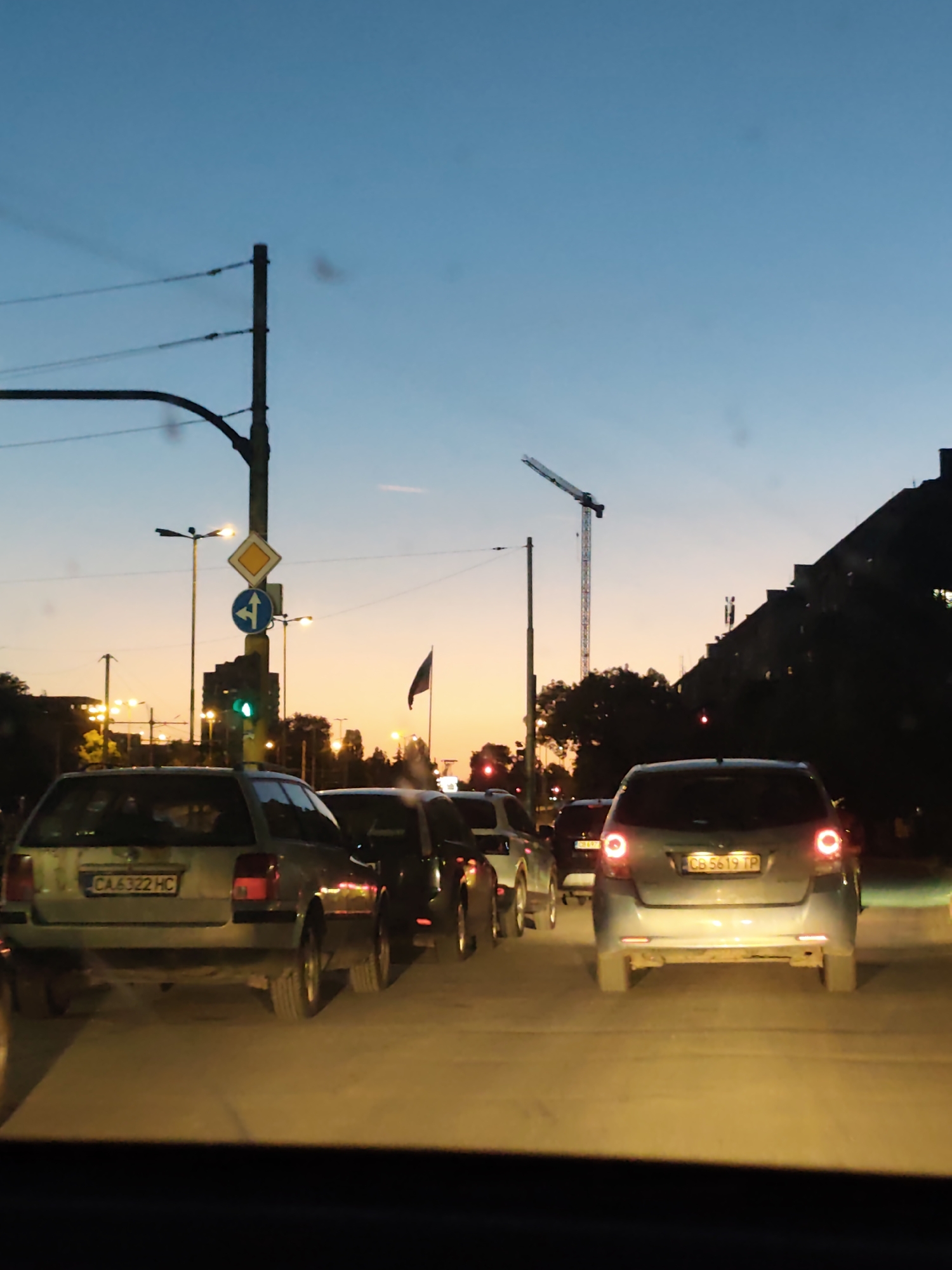 The image depicts heavy traffic at dusk in Sofia, Bulgaria. The sky transitions from blue to orange near the horizon. A construction crane is visible against the sky. Streetlights and traffic signals illuminate the scene. The image is taken from inside a car, looking out at the traffic ahead. Several cars are lined up, including a silver station wagon with license plate "CA6322 HC" and a small silver hatchback with license plate "CB 5619 TP." Road signs are present, including a directional sign with a roundabout arrow and a yield sign. A Bulgarian flag is flying in the background. Buildings line the street on the right side of the frame.
