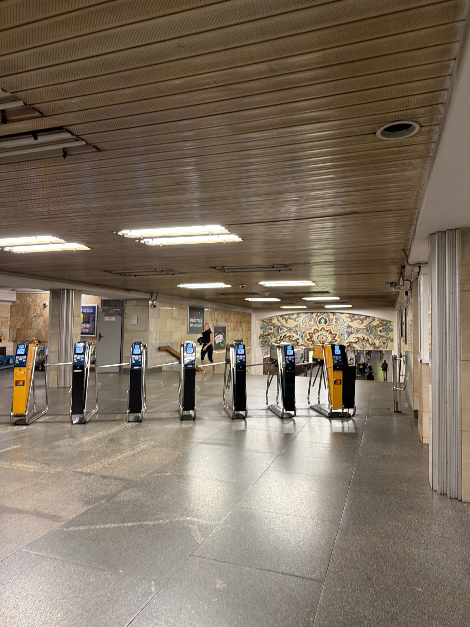 The image depicts the interior of a metro station in Kyiv, Ukraine. The scene is characterized by a long hallway with a tiled floor and a ribbed, beige-colored ceiling with fluorescent lighting. A series of ticket barriers, some yellow and black, others black, are positioned in the foreground, allowing passage through. On the left, a woman with dark hair and a black top is walking away from the camera, carrying a black bag. In the background, a colorful mosaic adorns the wall above another set of ticket barriers. To the right, a person is walking down the hallway, and further in the distance, several individuals are visible, some with luggage, suggesting a busy transit environment. The overall atmosphere is functional and utilitarian, typical of a metro station.