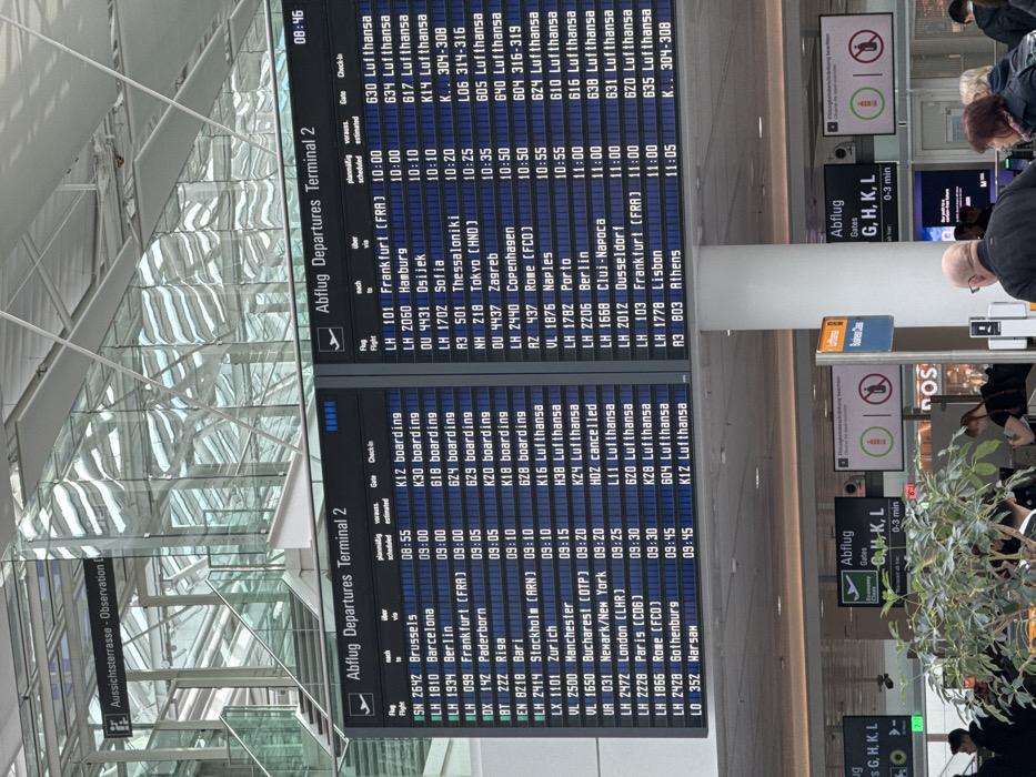 The image displays the interior of a modern airport terminal, identified as "Terminal 2 Abflug Departures," located at Munich Airport (Flughafen München Franz Josef Strauß) in Freising, Germany. Two large digital departure boards dominate the mid-ground, indicating the current time as 08:46 on the right board. These boards list various domestic and international flights, including destinations such as Brussels, Barcelona, Frankfurt, London, Paris, Hamburg, Tokyo, and Athens, with scheduled/estimated departure times ranging from 08:55 to 11:05, corresponding gates, and check-in statuses, with numerous flights identified as Lufthansa.
The terminal architecture features a high, vaulted ceiling constructed from a glass and metallic framework, allowing natural light into the space and creating reflections on the glass surfaces. Upper levels with glass railings and staircases are visible in the background, along with a sign pointing to "Aussichtsterrasse - Observation D." In the lower foreground, multiple individuals, predominantly adults, are present. A bald man in a dark jacket is centrally located in the lower right foreground, facing away from the camera. Other individuals are seen walking or standing, suggesting passenger movement. Green leafy plants in planters are positioned in the lower left and center foreground. Additional digital signs are visible, one indicating "Abflug Gates G, H, K, L 0-3 min" for Economy Class and another displaying "Business Class" with Lufthansa branding. Overhead signs feature iconography, including a crossed-out bottle, indicating restrictions. The scene depicts a functional and active airport environment during daylight hours.