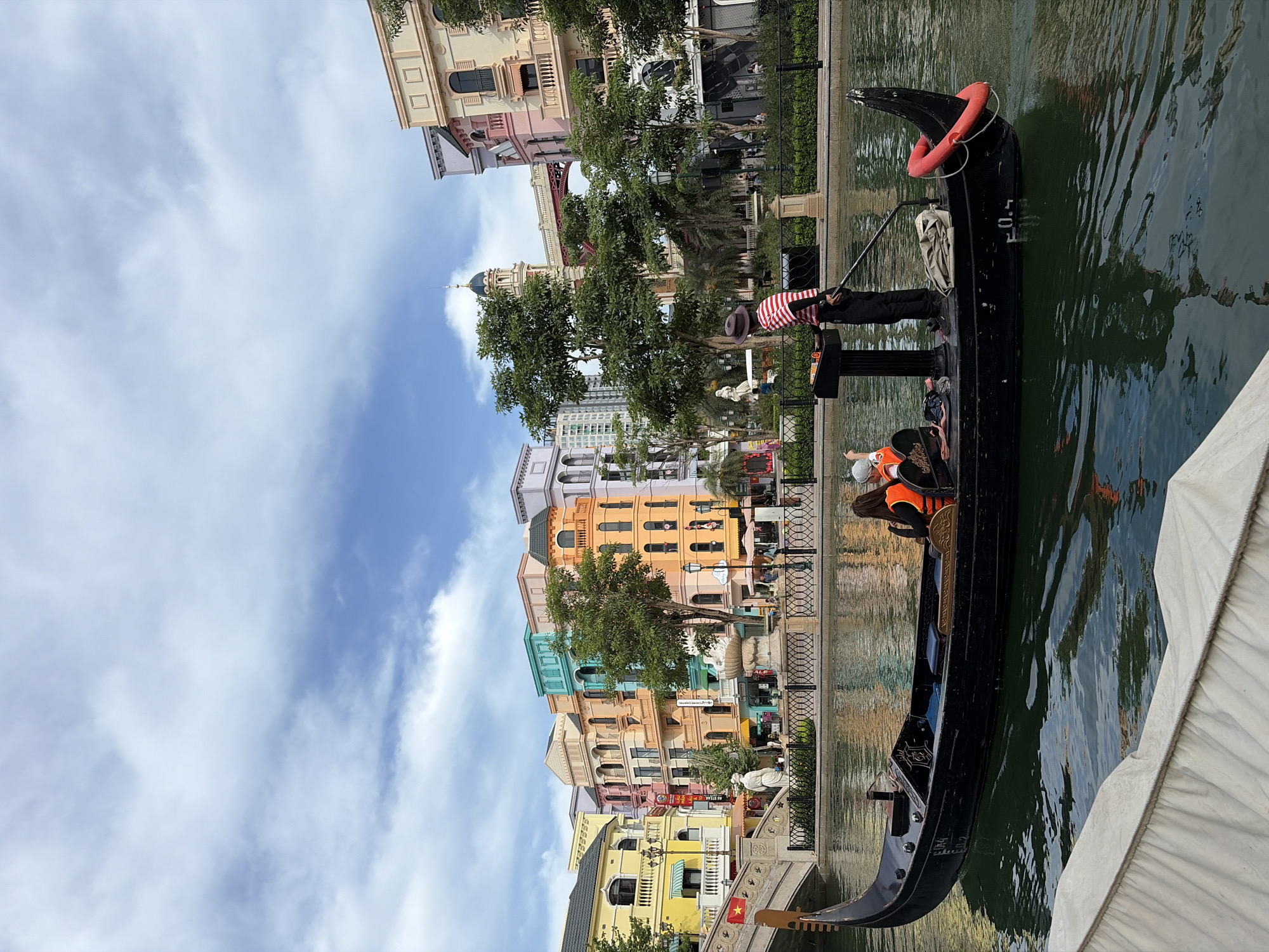 In the city of Nghĩa Trụ Commune, Vietnam, a black gondola navigates a canal lined with colorful buildings that resemble Venetian architecture. The buildings are painted in vibrant hues of yellow, pink, orange, and teal, featuring arched windows and ornate balconies. A gondolier, wearing a red and white striped shirt and a hat, stands at the stern of the gondola, holding a long oar. Two passengers, wearing orange life vests, are seated in the gondola. The canal water is calm and reflects the sky and buildings. In the background, a bridge arches over the canal, and trees with lush green foliage are interspersed among the buildings. The sky is partly cloudy with a bright blue expanse.