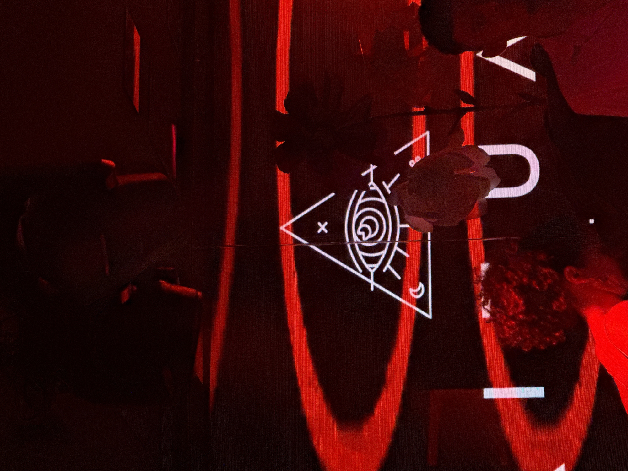The image shows a darkened interior space with a white symbol projected on a surface that appears to have red vertical accents. The symbol is a triangle containing an "x," a stylized eye-like design, and crescent moon-like shapes. Near the symbol is a blurred white "U" shape. Two blurred people are visible on the right side, one wearing red. Another blurry person is positioned on the upper left side of the image. There are shadows cast with a red hue, adding to the dimly lit atmosphere. There is no indication of the image location.