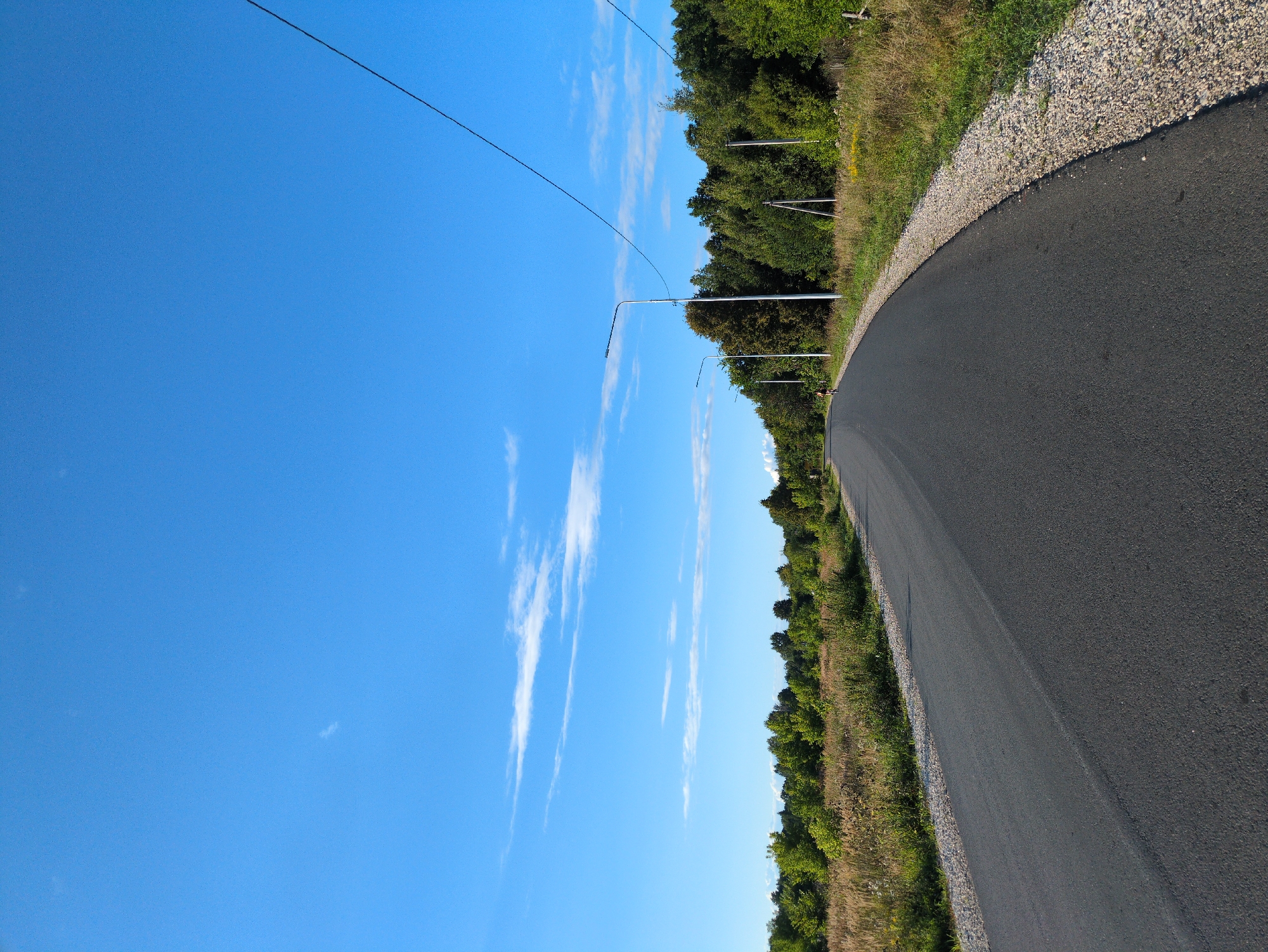 An asphalt road curves to the right through a rural landscape under a clear blue sky with scattered white clouds. The road is bordered by dry grass and vegetation on the left and a gravel shoulder and more vegetation on the right. A line of trees forms a dense backdrop along the right side of the road, with sparse trees and fields visible in the distance on the left. Several modern streetlights are spaced along the right side of the road. A single person is visible on a bicycle in the distance on the left side of the road. Electrical wires are strung across the upper right portion of the frame. The scene appears to be in a natural, undeveloped area, possibly on the outskirts of Kulebaki, Russia.