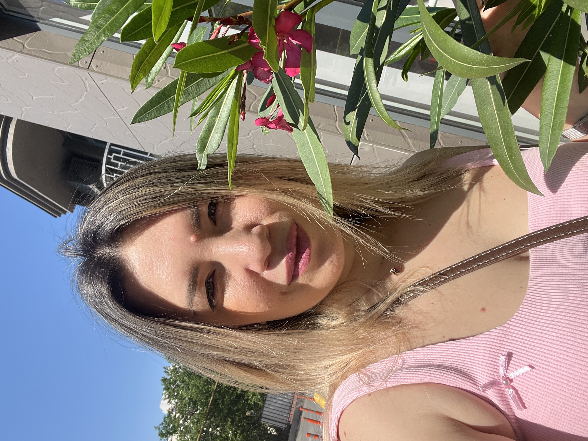 A woman is taking a selfie outdoors in Sochi, Russia, on a sunny day. She has blonde hair and is wearing a pink tank top. A branch with green leaves and pink flowers is in the foreground, partially obscuring her right side. In the background, a building with a modern architectural design and a balcony is visible. Trees and traffic cones can be seen further in the background.