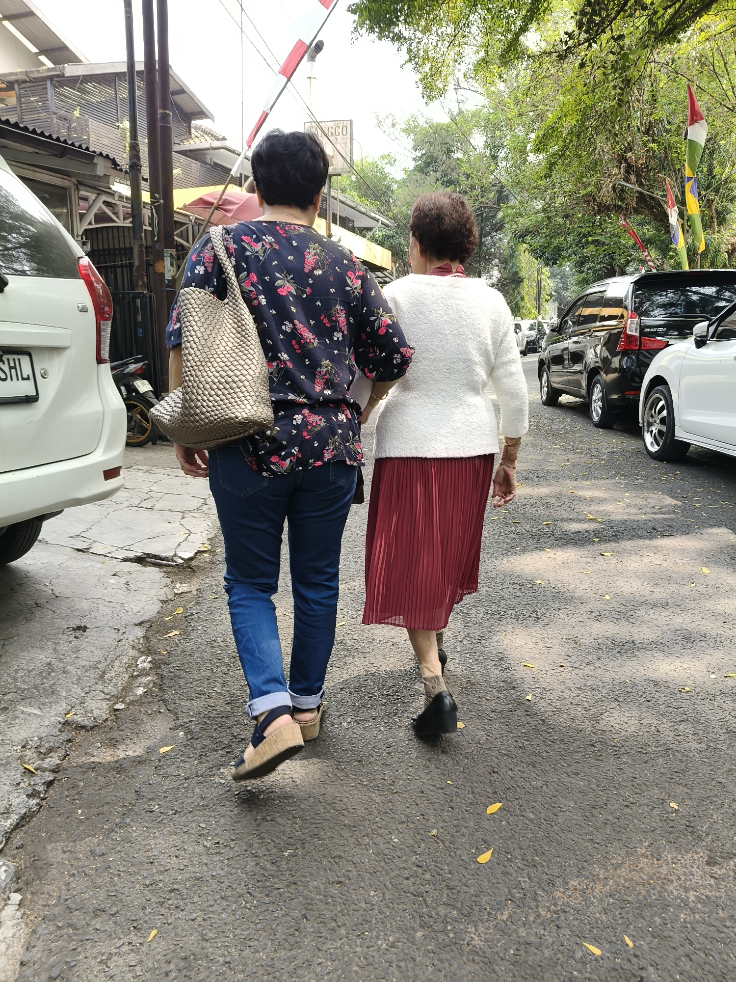 Two elderly women walk away from the camera down a paved road lined with trees and parked cars. The woman on the left wears a dark floral print shirt, blue jeans, and wedge sandals, carrying a large woven bag over her left shoulder. The woman on the right wears a white textured cardigan over a red pleated skirt and black heeled shoes. The scene appears to be in a residential or commercial area with buildings visible on the left side of the road. Flags are visible on poles in the background. The road surface is asphalt and scattered with fallen leaves. The image seems to have been taken in the city of Bandung, Indonesia, given the presence of parked vehicles and the general streetscape.