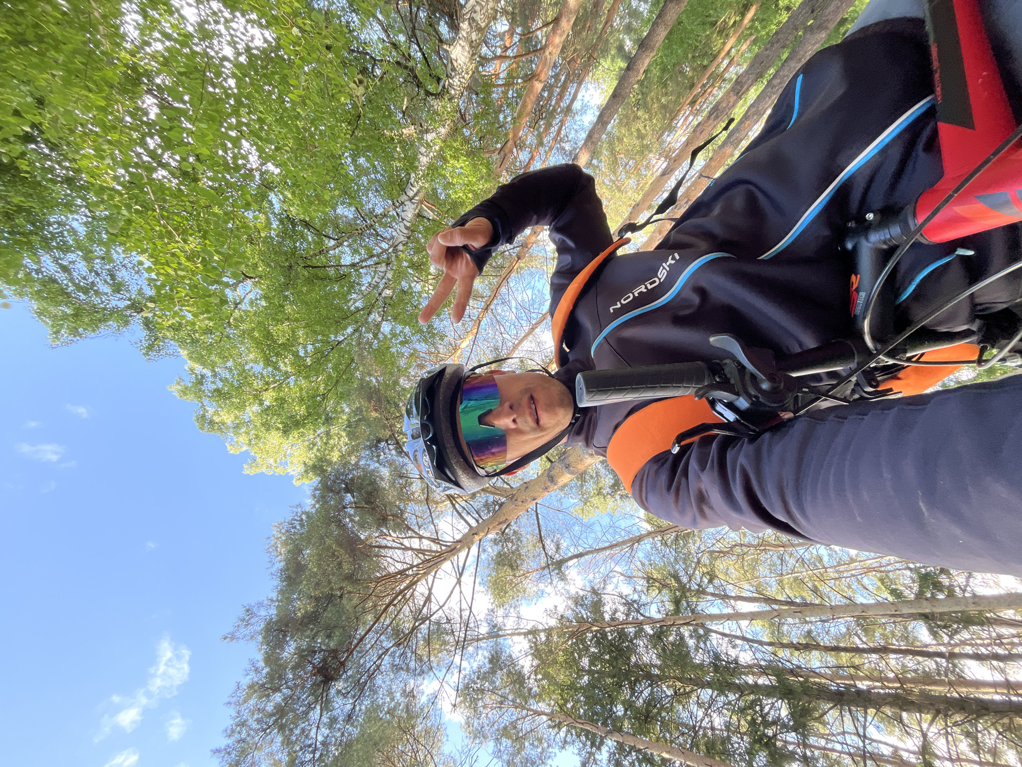 This upward-angled photograph captures a male cyclist in a forest setting, likely in Chaikovsky, Russia. The individual is taking a selfie, holding his left hand up in a "peace" sign, indicating a relaxed or positive mood.
He is equipped for cycling, wearing a dark blue long-sleeved jersey with light blue and white stripes, featuring the brand "NORDSKI" prominently on the shoulder. He also has on a black cycling helmet and distinctive iridescent, multi-colored reflective sunglasses. Parts of his bicycle are visible in the foreground, including the handlebars, a brake lever, and a red water bottle cage mounted on the frame. An orange sleeve or arm warmer is visible on his right arm.
The background showcases a dense forest canopy under a clear, bright blue sky with a few scattered white clouds. Sunlight filters through the leaves, suggesting a sunny late morning or early afternoon. The trees appear to be a mix of deciduous species with vibrant green foliage, possibly birch trees with their distinctive white bark, and coniferous trees with darker needles, consistent with the vegetation found in the specified region. The image conveys a sense of outdoor activity and connection with nature.