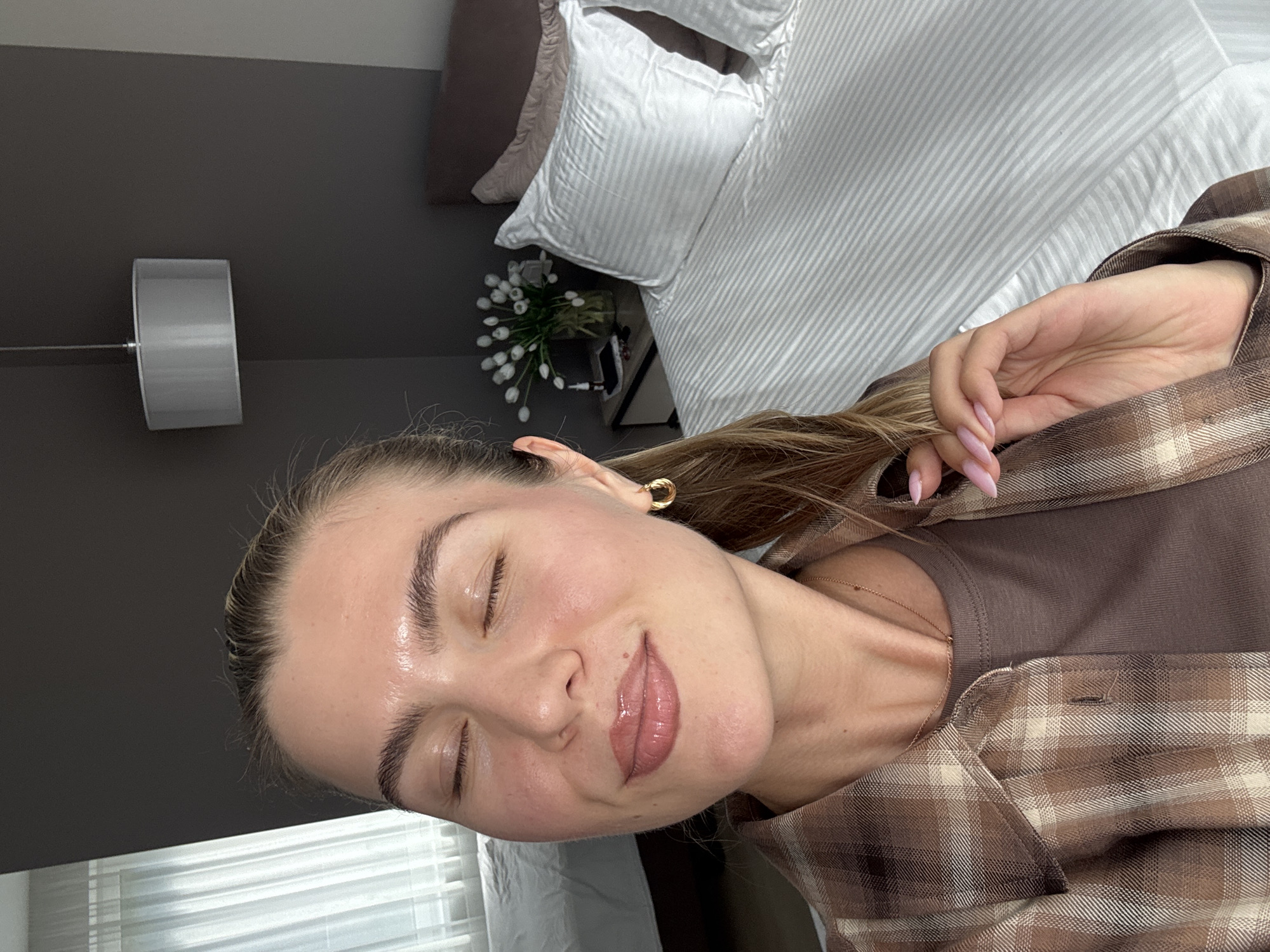 This is a close-up, head-and-shoulders portrait of a young woman indoors, possibly in a bedroom. The woman has her eyes closed and a slight smile on her face, appearing relaxed and content. Her skin is smooth and appears to have a dewy glow, and she is wearing lip gloss. Her eyebrows are dark and well-defined, and she has a gold hoop earring in her left ear. She is wearing a brown and cream plaid shirt over a brown t-shirt, and a delicate gold necklace. Her hair is long and light brown.
The background suggests an indoor setting, likely a bedroom. To the left, a dark grey wall is visible, with a cylindrical, grey lampshade mounted on it. Behind her, a neatly made bed with white, striped bedding and pillows is partially visible. A small vase with white flowers sits on a bedside table, adding a touch of decor. The lighting appears soft, suggesting it might be daytime, though no windows or direct light sources are clearly visible. The overall mood is peaceful and serene.