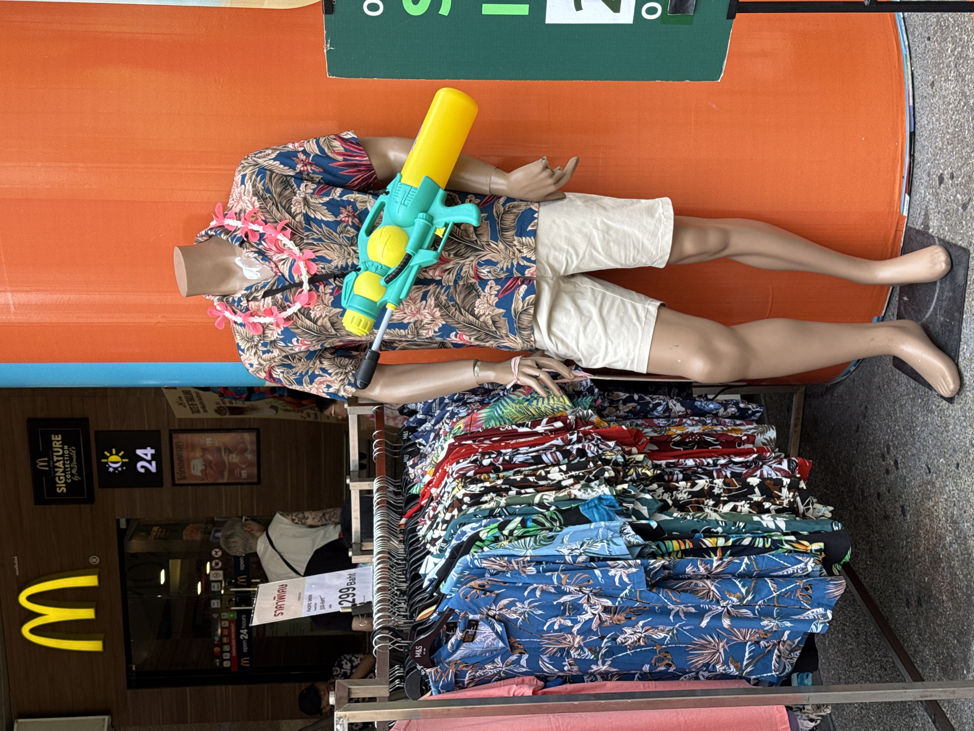 The image shows a mannequin holding a water gun, wearing a floral shirt, white shorts, and a lei, laid horizontally on top of a rack filled with folded floral shirts. The scene is in front of a McDonald's restaurant. A sign indicates that it is open 24 hours. The shirts are priced at 299 Baht. The location is likely in Bangkok, Thailand, given the presence of Baht currency. There is a person visible through the front of McDonald's, though mostly obscured, appearing to work inside the restaurant.