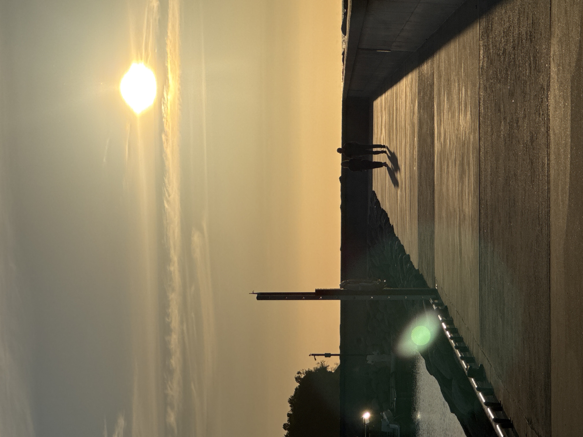 The image depicts a sunset scene on a pier in Tallinn, Estonia. The sun, a bright orb, is positioned in the upper right quadrant of the sky, casting a warm, golden glow over the horizon and the surrounding environment. The sky is a gradient of pale yellow to light orange, with wispy clouds streaked across it.
On the pier, two figures, silhouetted against the setting sun, are walking away from the camera. They are positioned in the middle ground, with their shadows stretching out before them on the paved walkway. The pier itself is constructed of concrete, with a darker concrete wall running along the right side. On the left side of the pier, a body of water is visible, with a rocky embankment lining its edge. In the distance, on the left, a treeline is discernible.
A metal pole with a life preserver attached stands on the left side of the pier, near the rocky embankment. Another, thinner pole is visible further in the background to the left. A green lens flare is present in the lower left portion of the image, originating from a light source not fully visible. The overall atmosphere is serene and tranquil, capturing the beauty of a sunset over the water.