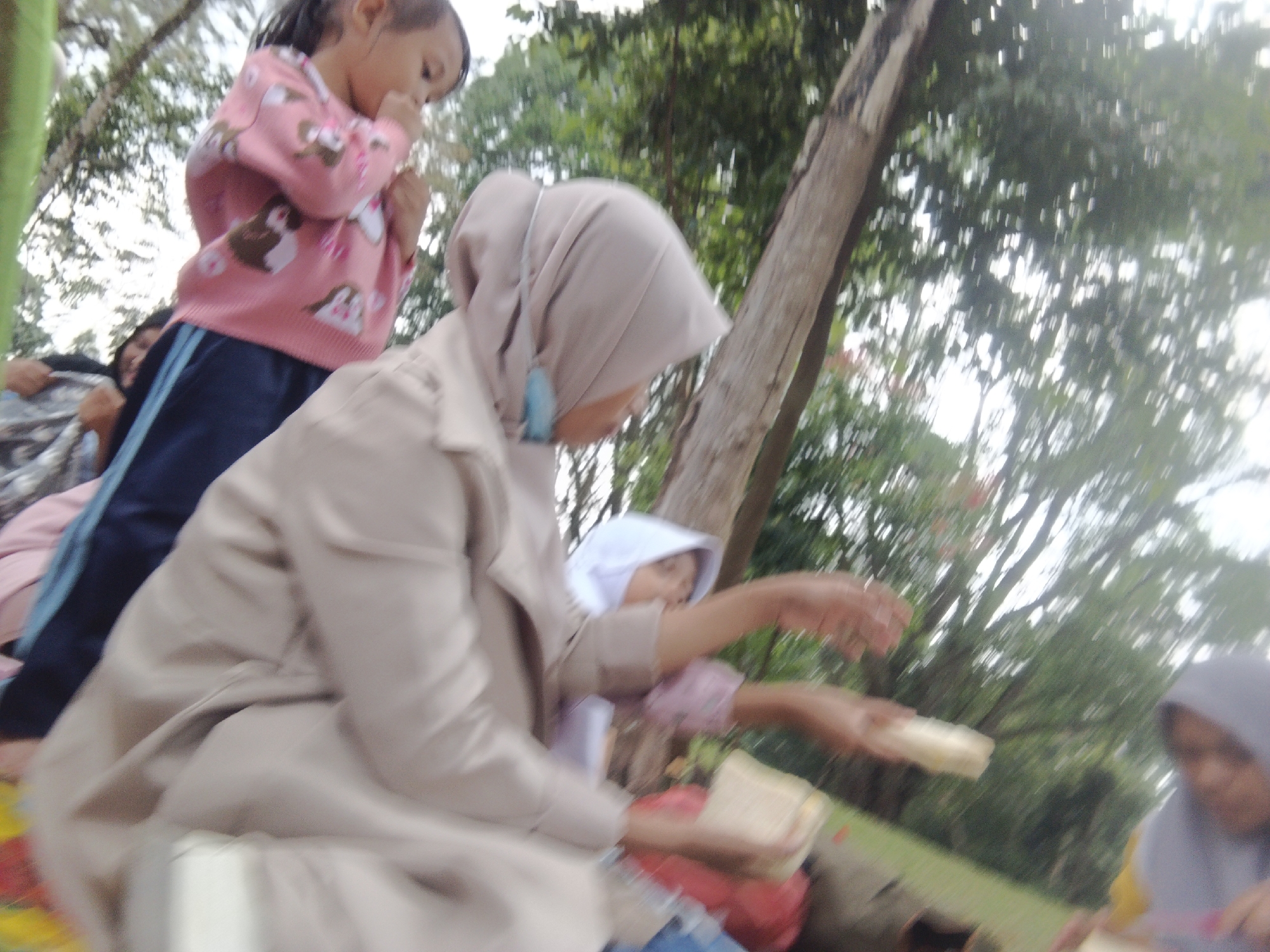 In Perbawati, Indonesia, on an outdoor grassy area beneath trees, a group of young girls is gathered. The girl in the foreground is wearing a light beige jacket and a headscarf. She is holding a light-colored object in her right hand. Next to her, another girl in a pink sweater with cartoon characters on it is visible from the waist up. She appears to be looking down and her hands are near her mouth. To the right, a younger child in a white head covering, possibly a younger sibling, is also present, with their arms outstretched. The background is filled with lush green trees and foliage, suggesting a park or natural setting. The image appears to be taken at a casual gathering, possibly a picnic or an outdoor activity.