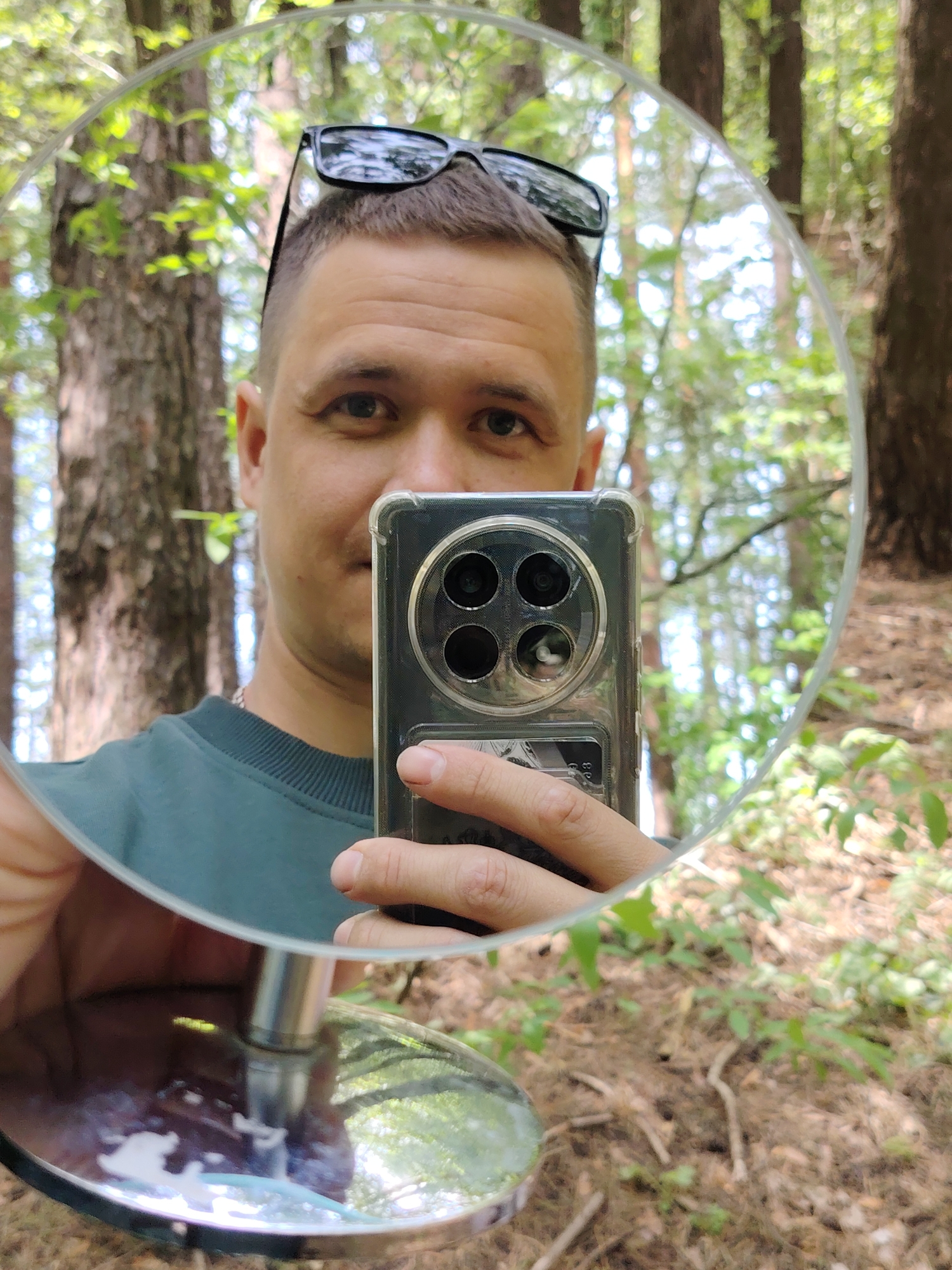 A man is holding a round mirror in his left hand, reflecting his face and a smartphone. The man has short brown hair, sunglasses on his head, and is wearing a dark green sweatshirt. He's holding a smartphone with a visible four-lens camera module in his right hand. The mirror is reflecting the surrounding environment, which appears to be a forest with trees and ground cover. The location is potentially a wooded area near Chaikovsky, Russia, based on the forest type and vegetation.