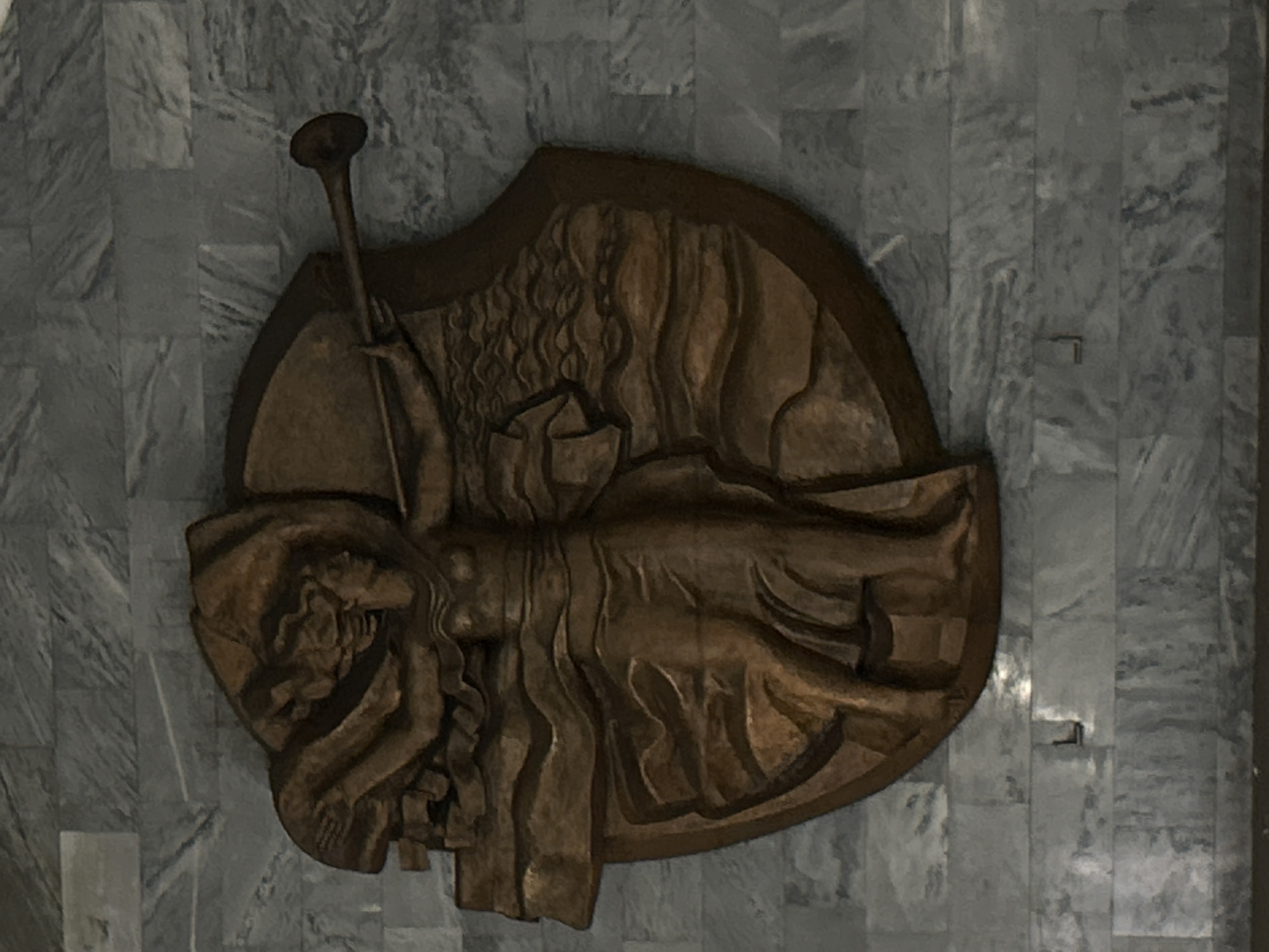 This media file captures a close-up of a bas-relief sculpture mounted on a tiled wall, likely in Saint Petersburg, Russia. The sculpture, rendered in a dark, rich brown, appears to be made of wood or bronze and features a stylized, elongated human figure, possibly female, in a reclining or supine pose.
The figure dominates the oval-shaped relief, positioned diagonally across the frame. It possesses flowing, voluminous hair and a draped garment, depicted with deep, undulating grooves that create a sense of movement and drama. The figure's left arm extends upwards, holding a long, slender staff or horn-like object with a flared end, suggesting an act of announcement or symbolic significance. The artistic style is expressive, with fluid lines and abstract forms, giving the piece a monumental yet ethereal quality.
The background wall consists of rectangular, greyish tiles with visible natural veining, characteristic of marble or polished stone, arranged in a grid pattern. The lighting is subdued, emphasizing the contours and shadows of the relief. There is no visible text in the image. The entire piece seems to be a decorative or commemorative element within an architectural interior, possibly a public building or cultural institution in Saint Petersburg.