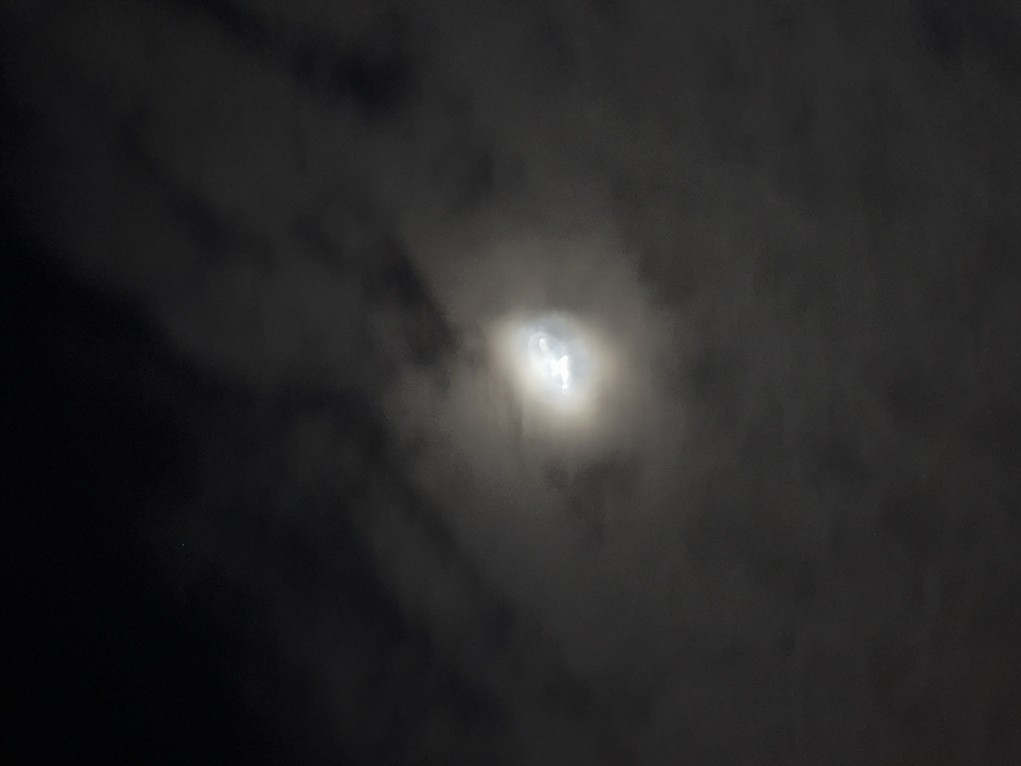 The image depicts a night scene, likely outdoors, with a bright celestial body partially obscured by clouds. The bright object, presumably the moon, shines through a layer of dark, diffuse clouds. The majority of the image is dark, transitioning from near-complete blackness in the lower left to varying shades of gray and white where the light from the moon interacts with the clouds. There is a small, bright point on the bottom left of the image, suggesting a star or light in the background. The location is given as Bac Nha Trang Ward, Vietnam.