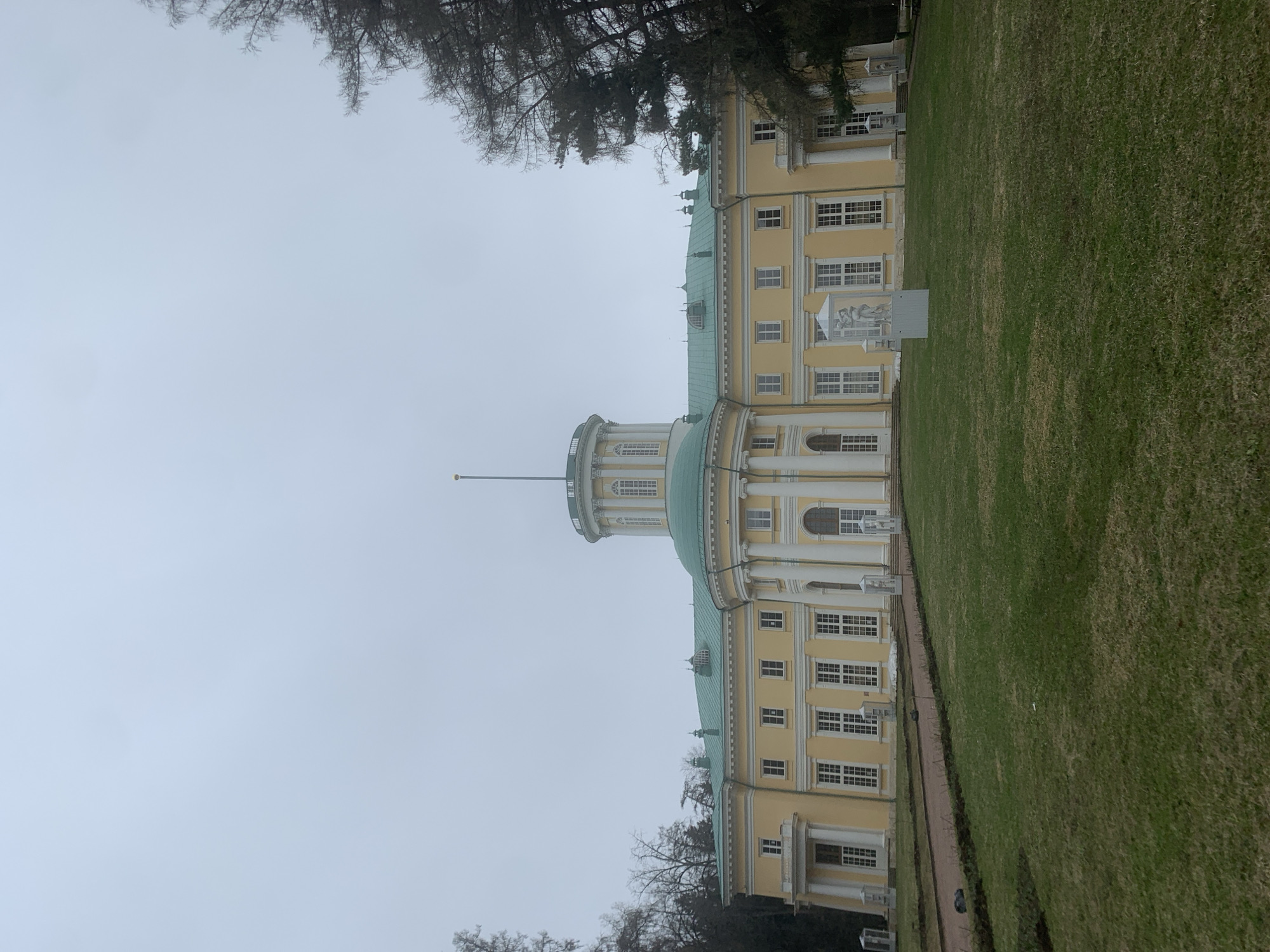 The image shows the main building of the Arkhangelskoye Estate in Russia. The building is predominantly yellow with white architectural detailing and a green roof. It features a central rotunda with a spire. The façade is lined with numerous windows, and there are decorative elements such as columns and statues. The building is surrounded by a large expanse of green lawn. Trees are visible on the left and in the foreground. The sky is overcast and grey. No people are clearly visible.