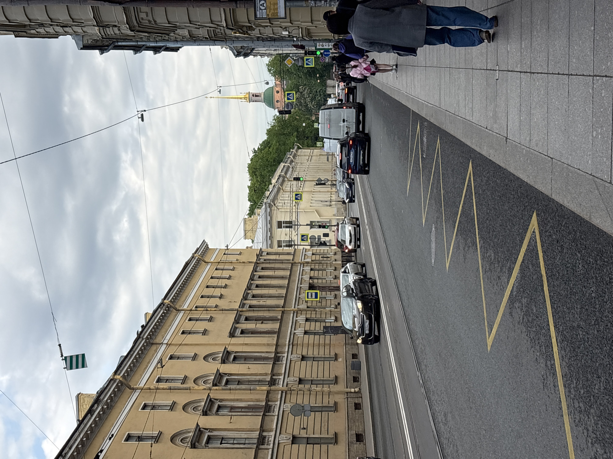 The image depicts a street scene in Saint Petersburg, Russia. The viewpoint is from the sidewalk looking down the street. On the right sidewalk, a few pedestrians are walking. A line of vehicles including cars and a van are traveling down the street. Markings including yellow zigzag lines are painted on the roadway. The architecture of the buildings lining the street is visible on the left, with a prominent building with a golden spire in the distance. Overhead electrical wires stretch across the sky. A street sign is attached to a building. The sky is overcast with cloudy skies.