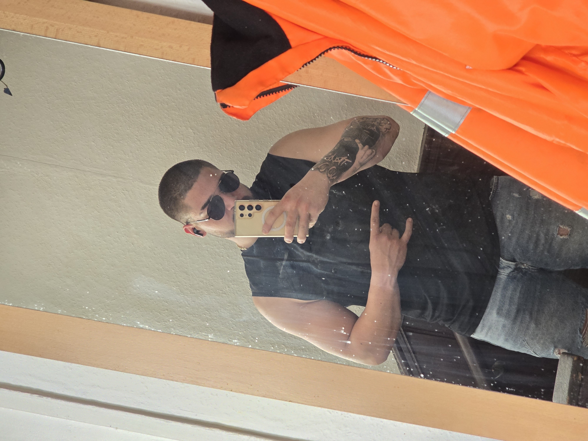 A man is taking a selfie in a mirror. He is wearing sunglasses, a black t-shirt, and distressed jeans. He has a tattoo on his left arm. He is holding a gold phone. An orange and black jacket is partially visible at the top of the frame. The background appears to be a room with white walls. The location in the city of Lutherstadt Eisleben, Germany is not discernible from the image.