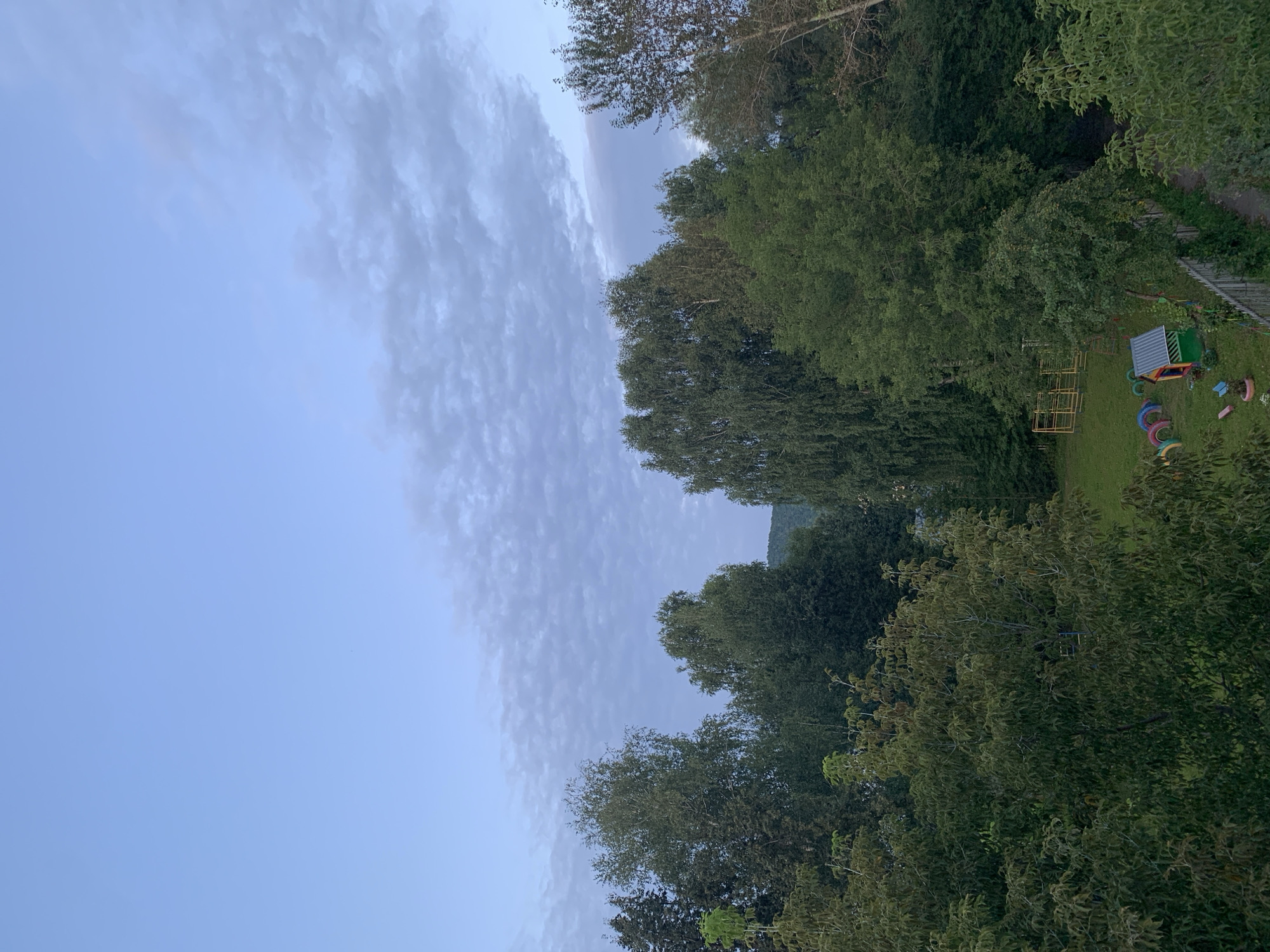 An overhead view captures a residential backyard in Asha, Russia, bathed in the soft light of early morning or late evening. The sky above is a pale blue, streaked with wispy clouds. Dominating the scene is a dense canopy of mature trees, their leaves a vibrant green, creating a natural border around the property.
In the lower right quadrant, a worn wooden fence delineates the edge of the yard. Beyond the fence, a dirt path meanders into the shadows. Within the yard, a small, colorful playhouse with a gray metal roof sits on the grass, its walls painted in bright red, yellow, and green. In front of the playhouse, a semicircle of repurposed tire swings, painted in various hues, lies on the ground. To the left of these, a child's slide, likely made of metal, is partially visible. Further back, a yellow metal playground frame, possibly for swings or a climbing structure, stands on the grassy lawn. The overall impression is one of a tranquil, somewhat rustic, domestic setting.
