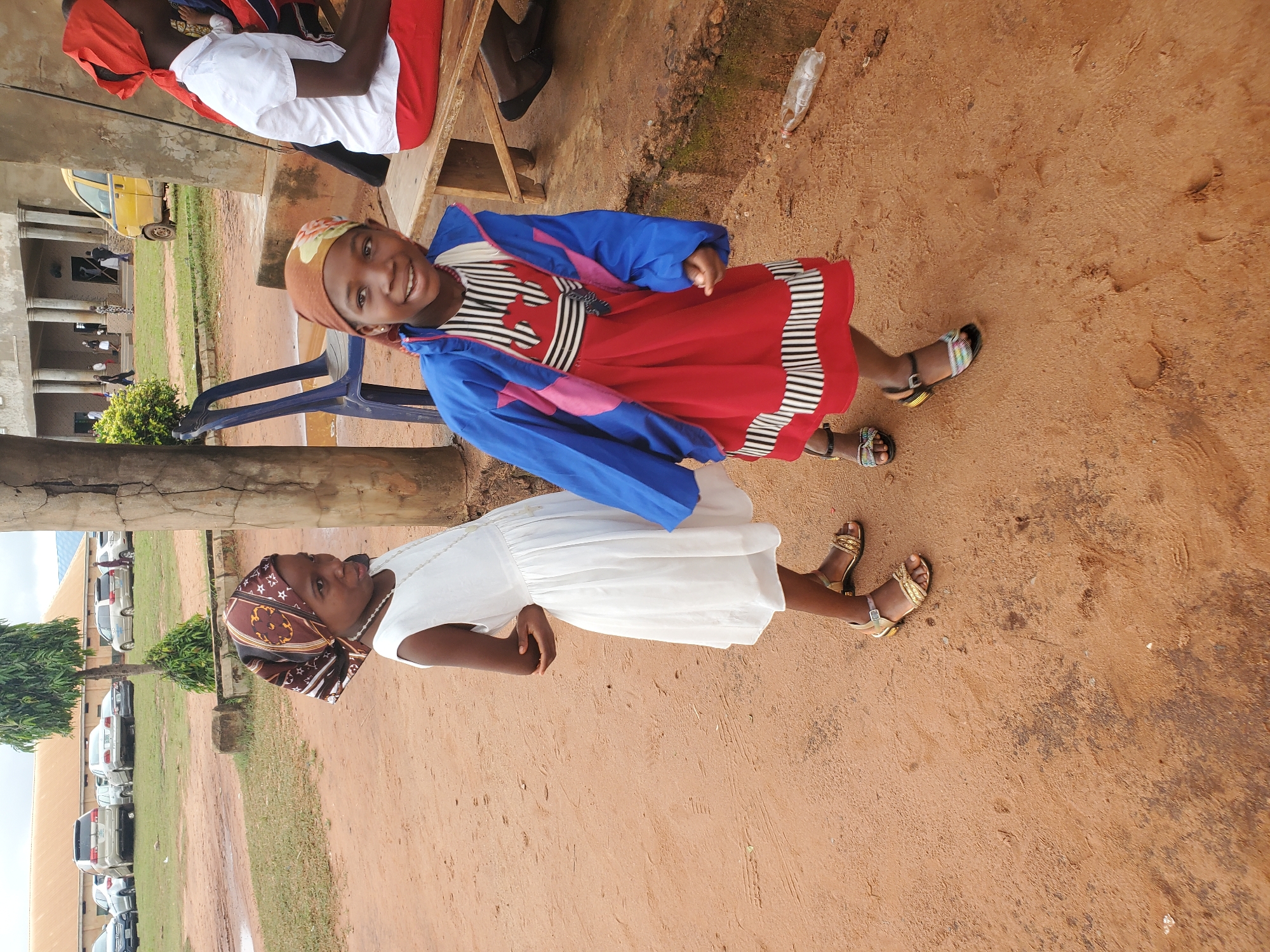 The image captures a scene in Nsugpe, Nigeria, under an overcast sky. Two young girls are the primary subjects, standing on reddish-brown dirt. The girl on the left wears a white dress and a patterned headscarf, with her arms crossed. The girl on the right is more animated, smiling and looking towards the camera, dressed in a red dress with black and white striped accents, a blue jacket, and a colorful headscarf. She wears a beaded necklace.
Behind the girls, the environment includes a large, light-colored building with columns, suggestive of a school or community center, and parked vehicles, including a yellow van. A blue plastic chair and a wooden bench are visible, along with discarded items like a plastic bottle. The ground is a mix of dirt and some wet patches. The overall impression is of an outdoor gathering or event in a semi-rural Nigerian setting.