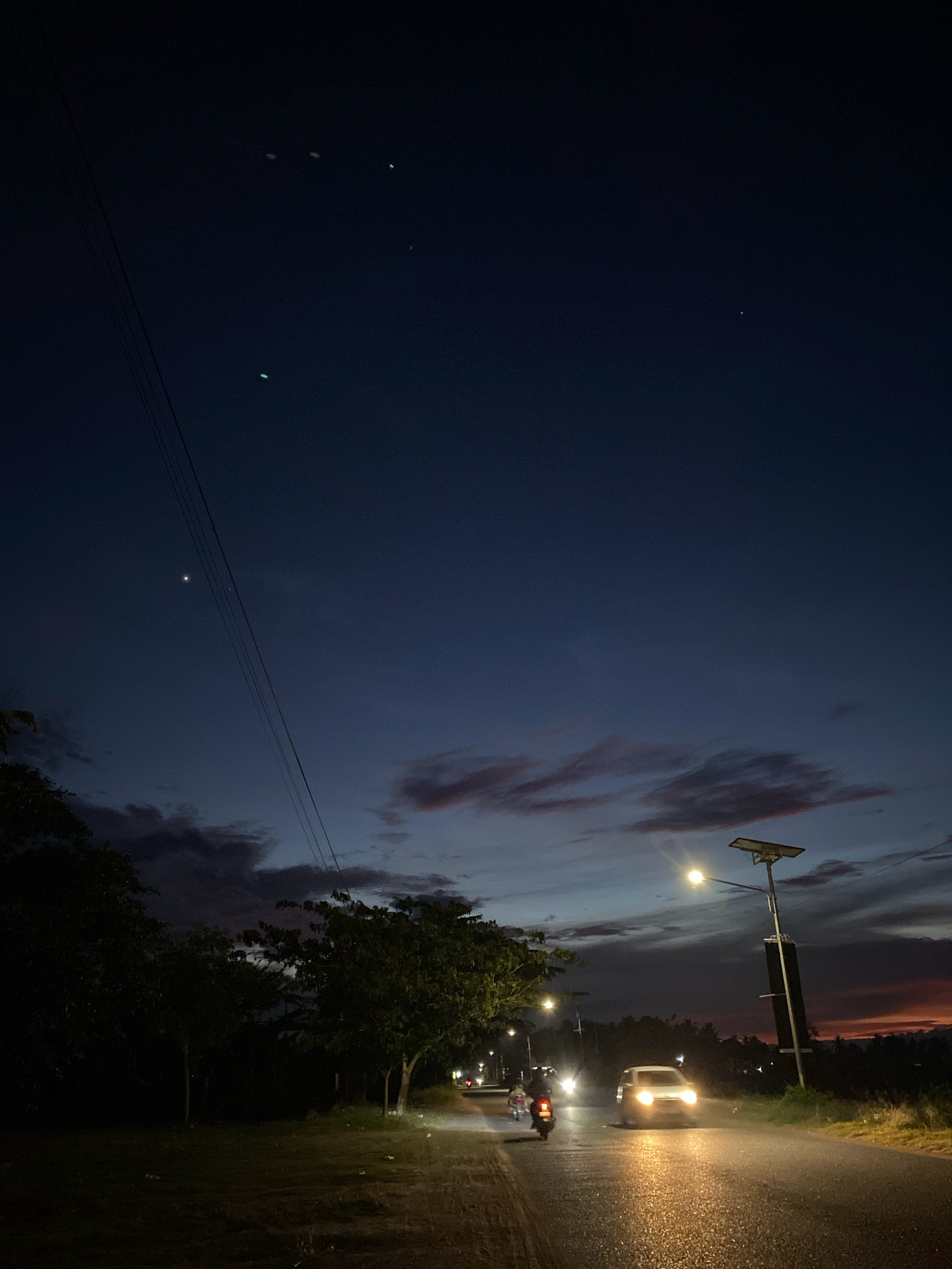 The image depicts a twilight scene on a road in Cotgapu, Indonesia. The sky is a deep blue transitioning to hues of purple and orange near the horizon, with scattered clouds. Power lines stretch diagonally across the upper left portion of the frame. In the foreground, a paved road is wet, reflecting the lights of passing vehicles and street lamps.
Several vehicles are visible on the road. A white car with its headlights on is moving towards the viewer. To its left, a motorcycle with a rider is also traveling in the same direction. Further down the road, other vehicles and their lights are visible. Streetlights, some equipped with solar panels, illuminate sections of the road.
On the left side of the road, there is a grassy area with trees, some of which are silhouetted against the sky. The overall atmosphere is that of an evening commute or travel on a rural or semi-rural road in Indonesia.