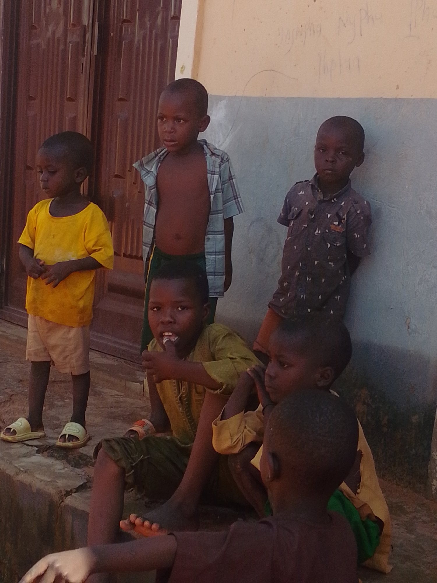 In the city of Tempure, Nigeria, a group of six young children are gathered outside a building. The building features a corrugated metal door on the left and a light-colored wall with some writing on it to the right. The children are of varying ages and are dressed in casual clothing.
On the far left, a young boy in a bright yellow t-shirt and light-colored shorts stands facing left, looking down. To his right, a slightly older boy in a plaid shirt, worn open, is also looking down. Behind him and leaning against the wall is another boy wearing a dark patterned shirt.
In the foreground, three children are seated. The one in the center, wearing a yellowish-green outfit, is looking at the camera and has his finger in his mouth. To his right, a boy in a yellow shirt with green fabric underneath is also seated. The youngest child in the group is partially visible in the lower right corner, facing away from the camera. The ground appears to be a mix of dirt and rough concrete.