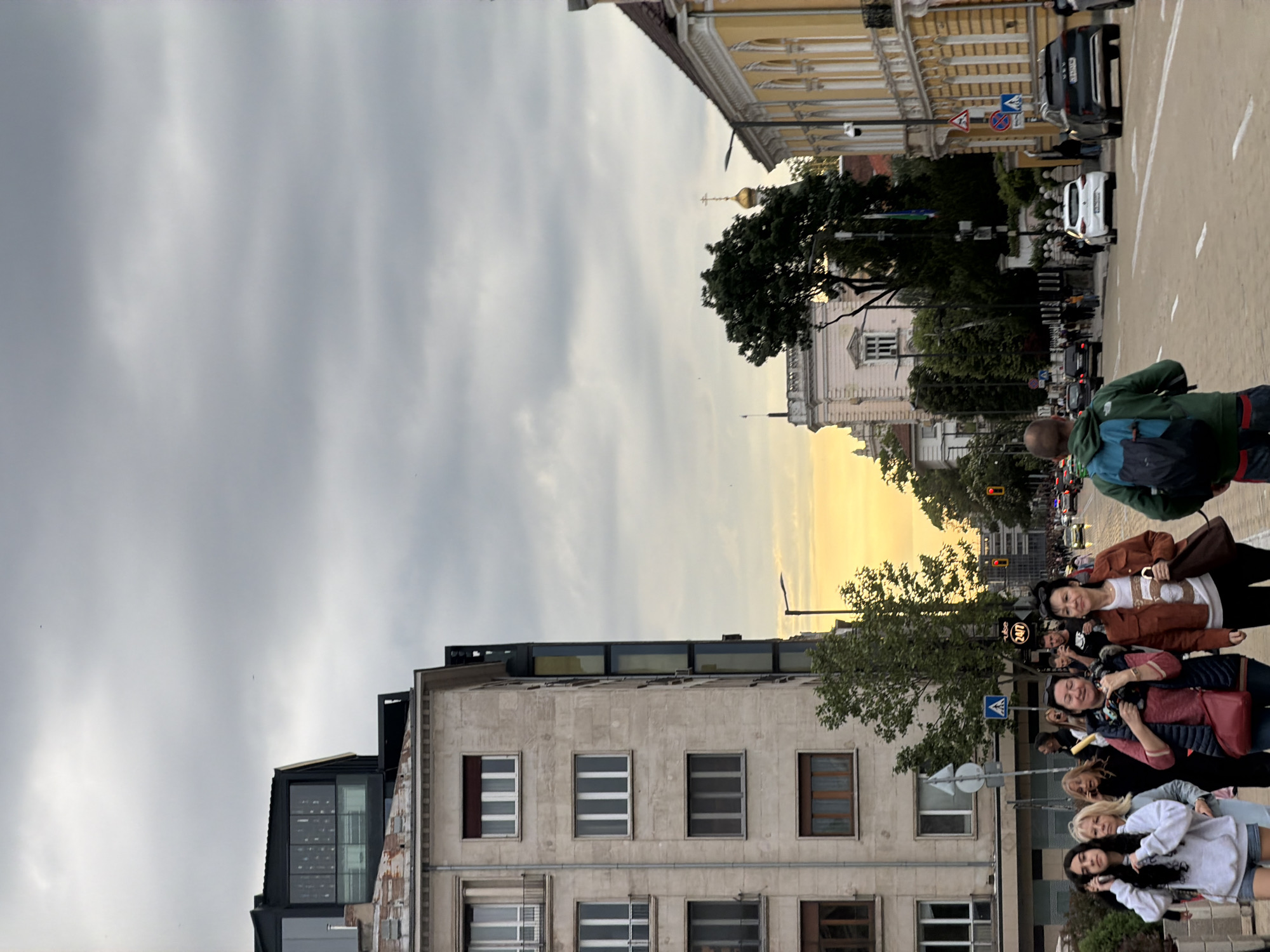 The image depicts a street scene in Sofia, Bulgaria. A group of women stand on the sidewalk along a building with windows. A man with a backpack walks on the street toward the camera. Cars are parked and moving along the street. Street signs are visible. The sky is cloudy. A building with a gold dome is in the distance. Buildings line the street, indicating an urban environment.