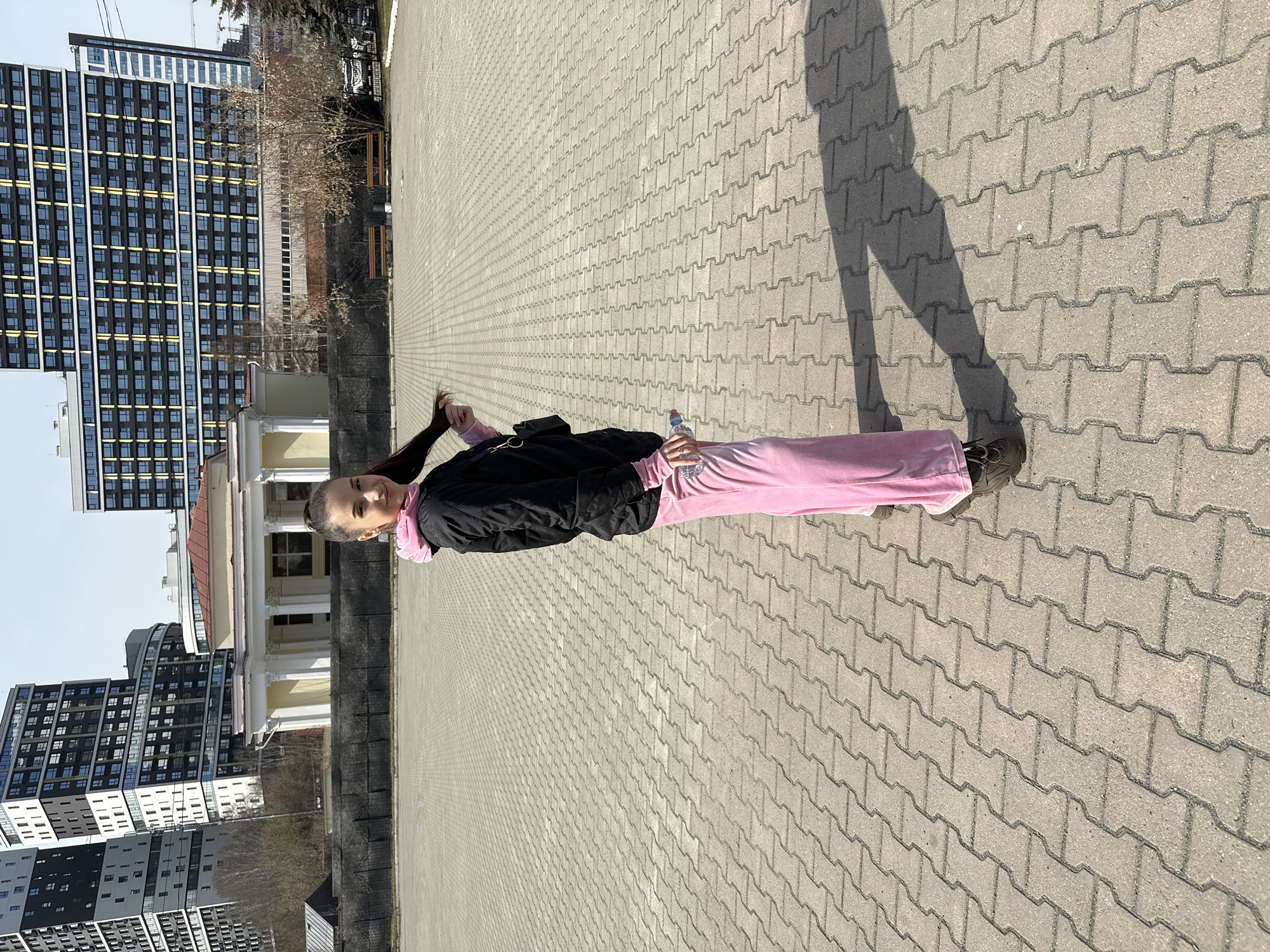 A woman stands in a large paved area in Yekaterinburg, Russia. She wears a black jacket, pink pants, and dark shoes, holding a water bottle in her right hand and touching her hair with her left. A long shadow stretches behind her on the paving stones. In the background are buildings, including a modern high-rise apartment building, as well as a building with white columns and a red roof. The day appears bright and sunny.