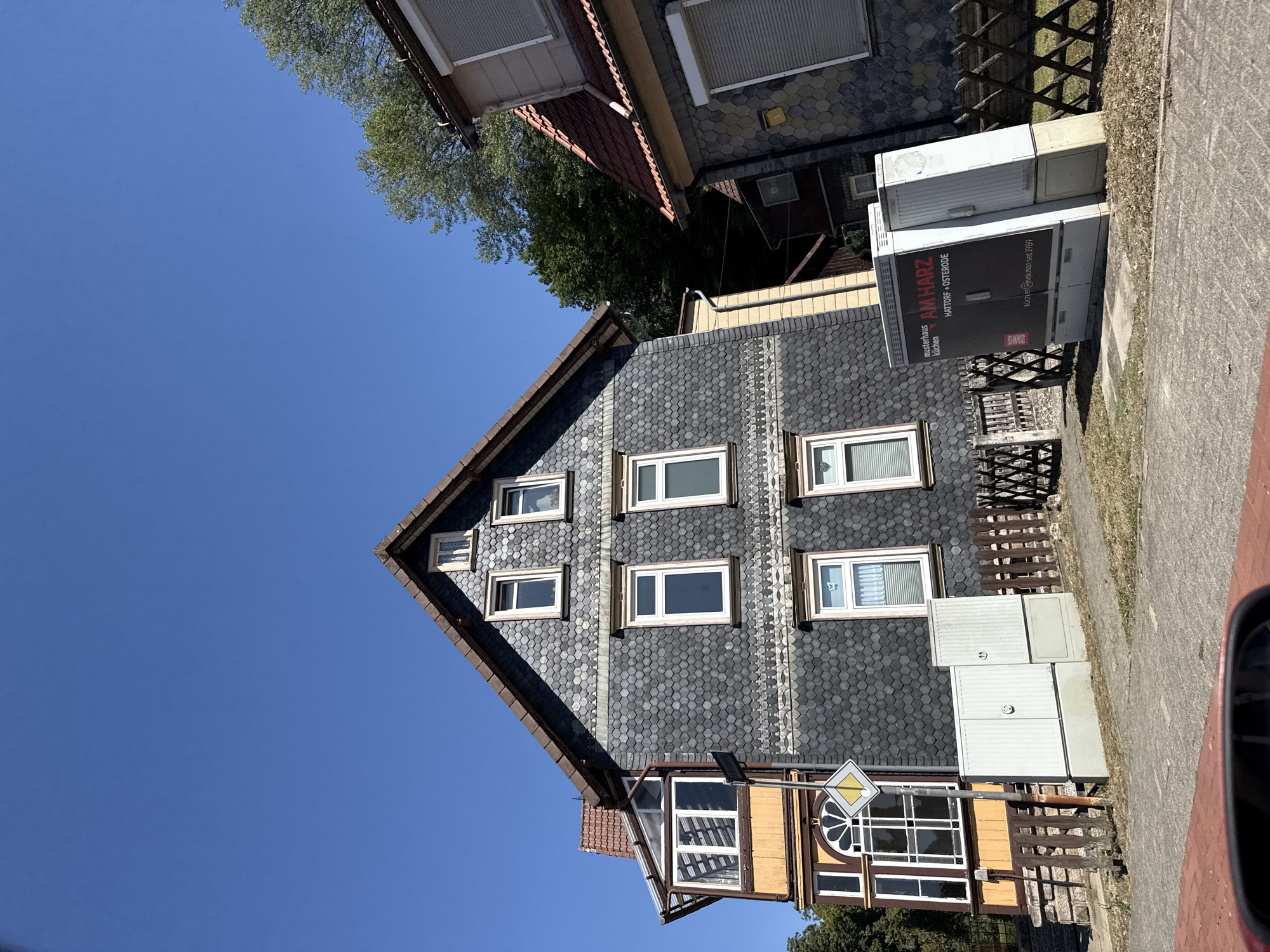 The image captures a street view in Bad Lauterberg im Harz, Germany. On the left, a multi-story house with a dark, hexagonal-tiled facade and a red-tiled roof dominates the scene. It features a bay window with a yellow-painted lower section and a glass enclosure above. Several windows with white frames are visible on the facade, some with decorative elements. In front of the house, there is a wooden fence, a sidewalk, and a paved road. Two white utility boxes are situated on the sidewalk, one of which has a black advertisement panel with text in German, including "AM HARZ" and "KÜCHEN REVOLUTION SEIT 1989". A traffic sign with a yellow diamond shape is mounted on a pole. To the right of the main house, another building with a similar tiled facade and a red roof is partially visible. Trees are present in the background, suggesting a natural environment surrounding the urban area. The sky is clear and blue, indicating a sunny day. No people are present in the image.