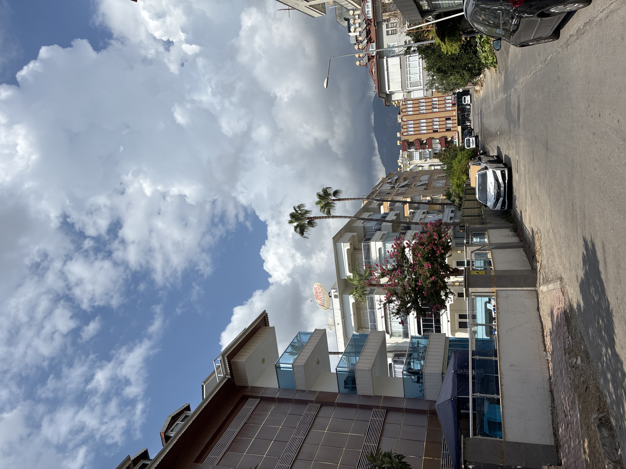 The image depicts a street scene in Alanya, Turkey, on a cloudy day. In the foreground, a paved road runs through a residential area lined with multi-story buildings. Several cars are parked along the street. A dark-colored tent covers an outdoor seating area in front of a building on the left. A flowering tree with pink blossoms is visible near the center of the image. In the background, palm trees and mountains can be seen. The sky is filled with large, fluffy white clouds. The architecture suggests a Mediterranean style, with balconies and awnings. The overall impression is one of a typical urban environment in a warm climate.