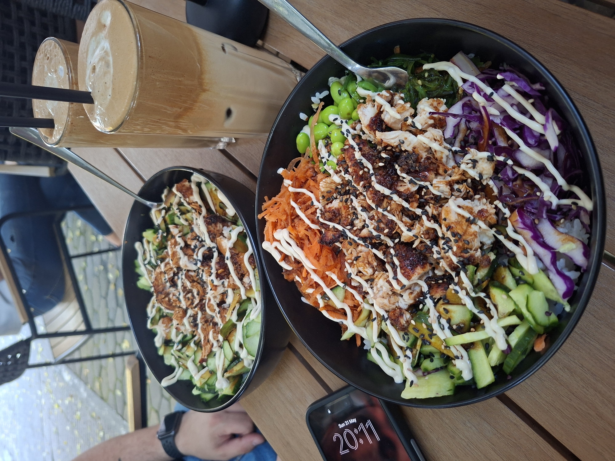 The image shows two black bowls of what appear to be poke bowls, likely taken at a restaurant or cafe in Plovdiv, Bulgaria. The bowls contain ingredients like rice, shredded carrots, cucumbers, red cabbage, edamame, seaweed salad, some kind of protein (possibly grilled chicken or fish), and a white sauce drizzled on top, along with black sesame seeds. Two tall glasses of iced coffee with straws are also visible. A smartphone displaying a time of 20:11 on Sunday, May 31, rests near the larger bowl. The scene takes place on a wooden table, perhaps in an outdoor setting. A watch is seen on the wrist of an individual on the bottom left. The lighting suggests daylight.