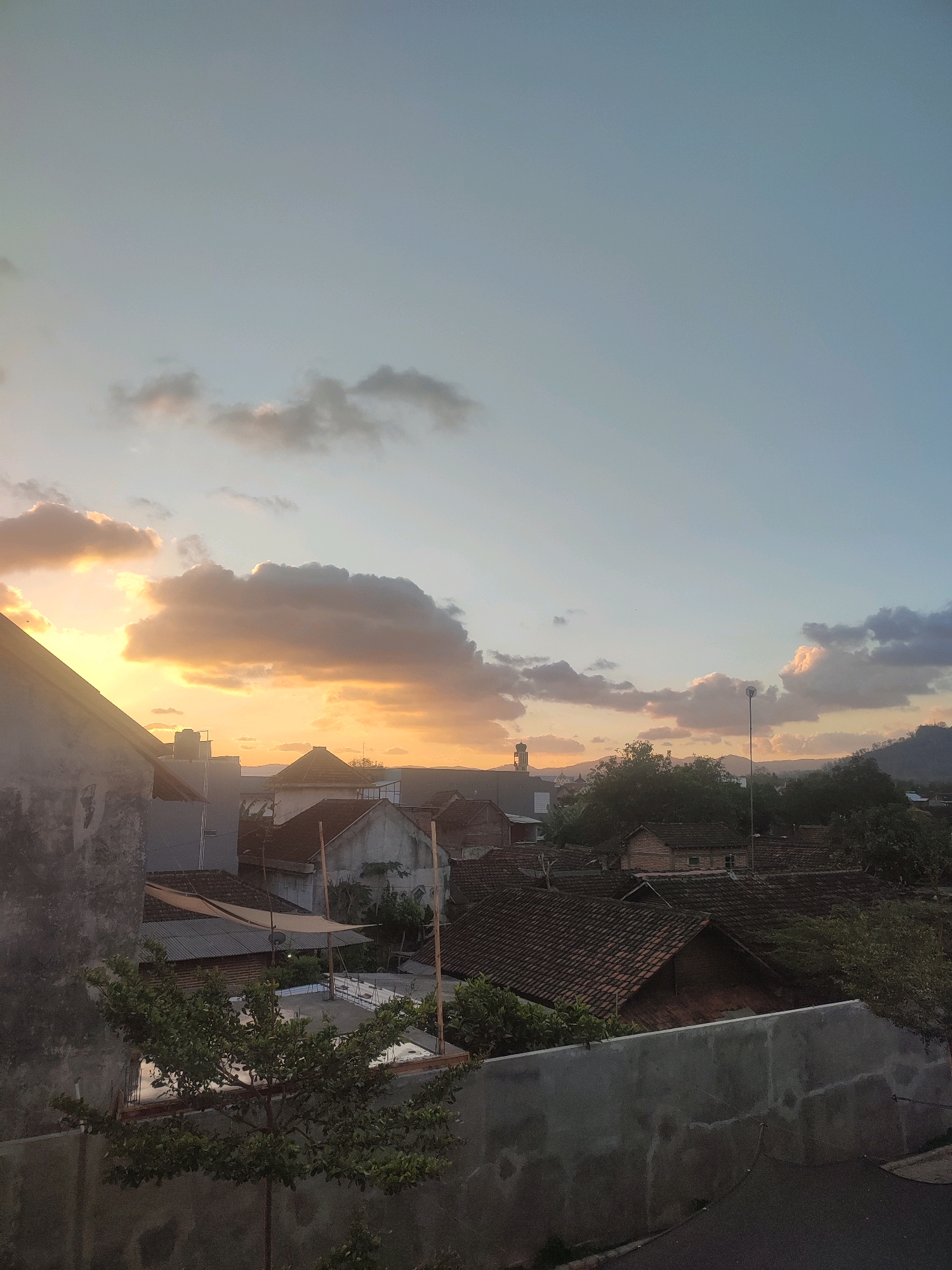 The image captures an outdoor scene in the city of Trenggalek, Indonesia, during what appears to be late afternoon or early evening. The sky is a gradient of pale blue and light orange, with scattered clouds reflecting the warm tones of the setting sun. The sun itself is not directly visible but its presence is indicated by the bright illumination on the clouds and the soft light casting across the landscape.
In the foreground, a textured concrete wall runs horizontally, with a dark asphalt road or pathway visible below it on the right. A small, leafy tree grows in front of the wall on the left. Behind the wall, the rooftops of several houses are visible, mostly covered with terracotta tiles. The architecture suggests a residential area. Some houses have visible walls made of rough plaster or concrete.
Further back, more houses and buildings are nestled amongst dense green trees. A water tank is visible on the roof of one building. In the distance, rolling hills or mountains form the horizon line. A tall, slender structure resembling a minaret or a tower is visible to the left of the center, with a few other distant buildings. The overall impression is a peaceful, semi-urban landscape bathed in the warm glow of the sunset. There are no people visible in the image.