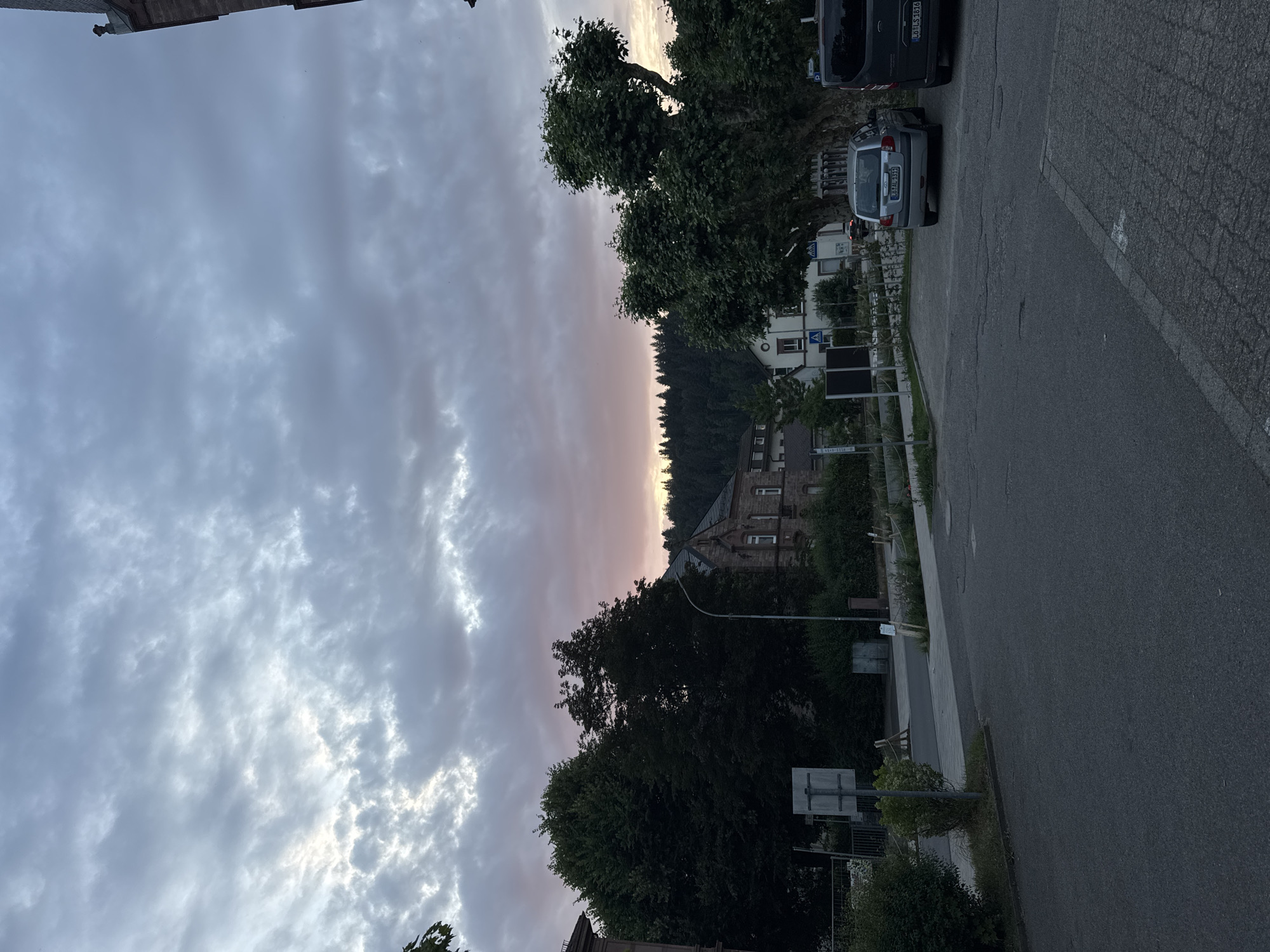 The image depicts a street scene in Schopfheim, Germany, during twilight. The sky is filled with dramatic, cloudy formations tinged with pink and orange hues, indicating the setting sun. In the foreground, a paved road and sidewalk are visible. To the right, two vehicles are parked: a silver hatchback and the rear of a dark van. Several trees with dense green foliage are present, with one particularly large, mature tree dominating the right side of the frame. Behind the trees, buildings with traditional architecture can be seen. A stone building with multiple windows and a pitched roof stands prominently. In the distance, a dense forest covers a hillside. The overall atmosphere is quiet and serene, capturing the transition from day to night.