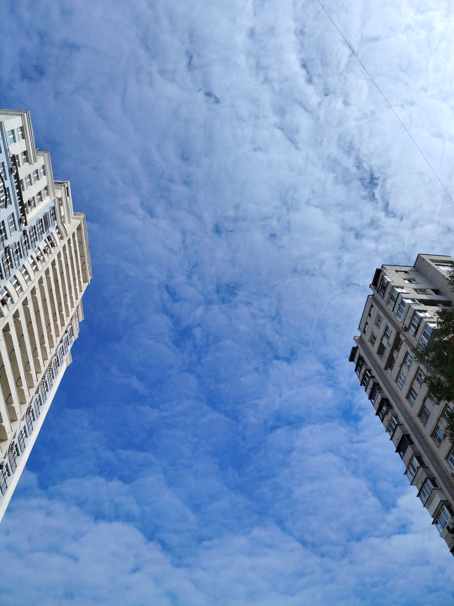 This low-angle, wide shot captures two apartment buildings against a bright blue sky with scattered white clouds. The building on the left is a modern, multi-story structure with a cream-colored facade and balconies. The building on the right, appearing older, is constructed from grey brick and also features balconies. Power lines stretch across the sky between the two structures. The image was taken in Kyiv, Ukraine. There are no people visible in the scene.