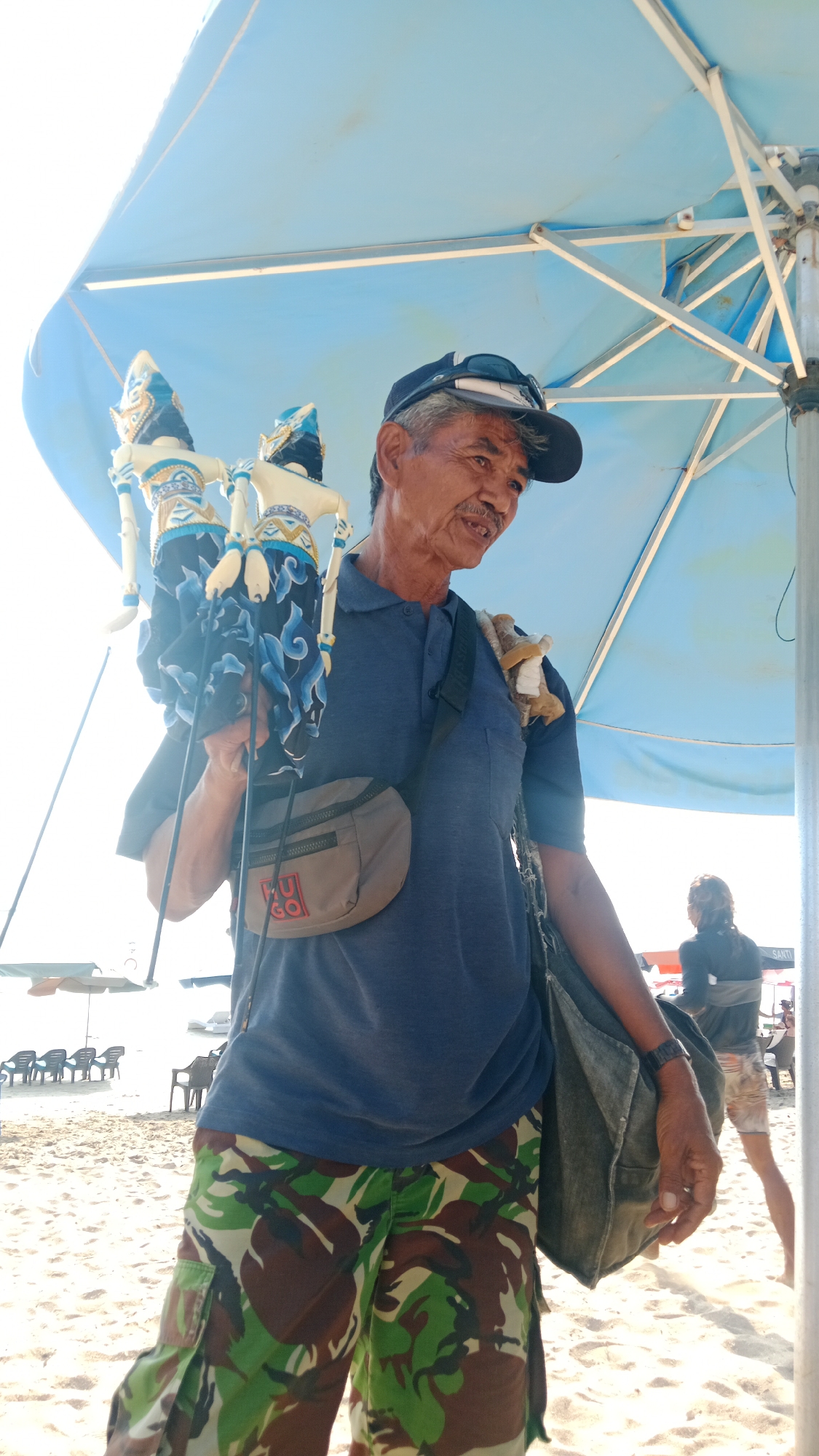 A man stands on a sandy beach, holding two wayang puppets under a large, light blue umbrella. He wears a dark blue polo shirt, a camouflage-patterned knee-length skirt, a dark baseball cap, and sunglasses. A small, tan waist bag with a red logo is slung across his front, and a large, dark green shoulder bag hangs on his right side. The puppets, dressed in white and blue traditional attire, are held by their strings. In the background, several empty beach chairs are visible on the sand, and a person in athletic wear walks away from the camera. The scene is brightly lit by sunlight.