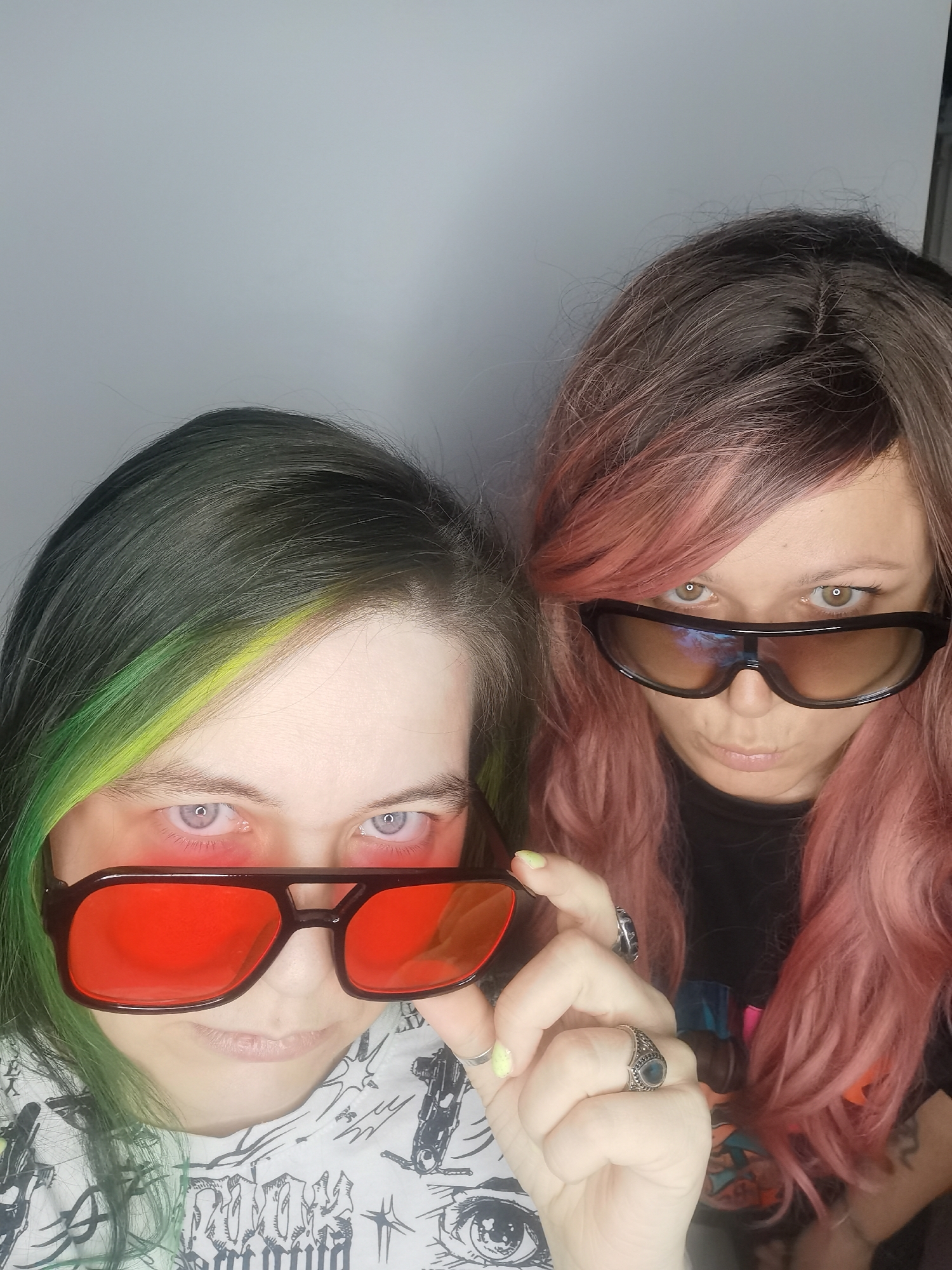 Two women stand close together, gazing directly at the camera. The woman on the left has green and yellow dyed hair and is wearing red-tinted sunglasses. She is holding the glasses with her right hand, revealing her purple eyes. She wears a white t-shirt with a gothic-style print. The woman on the right has brown and pink dyed hair and wears dark brown sunglasses. She is dressed in a black t-shirt. Both have a serious expression. The background is plain white. There is no indication that the image was taken in the city of Chaikovsky, Russia.