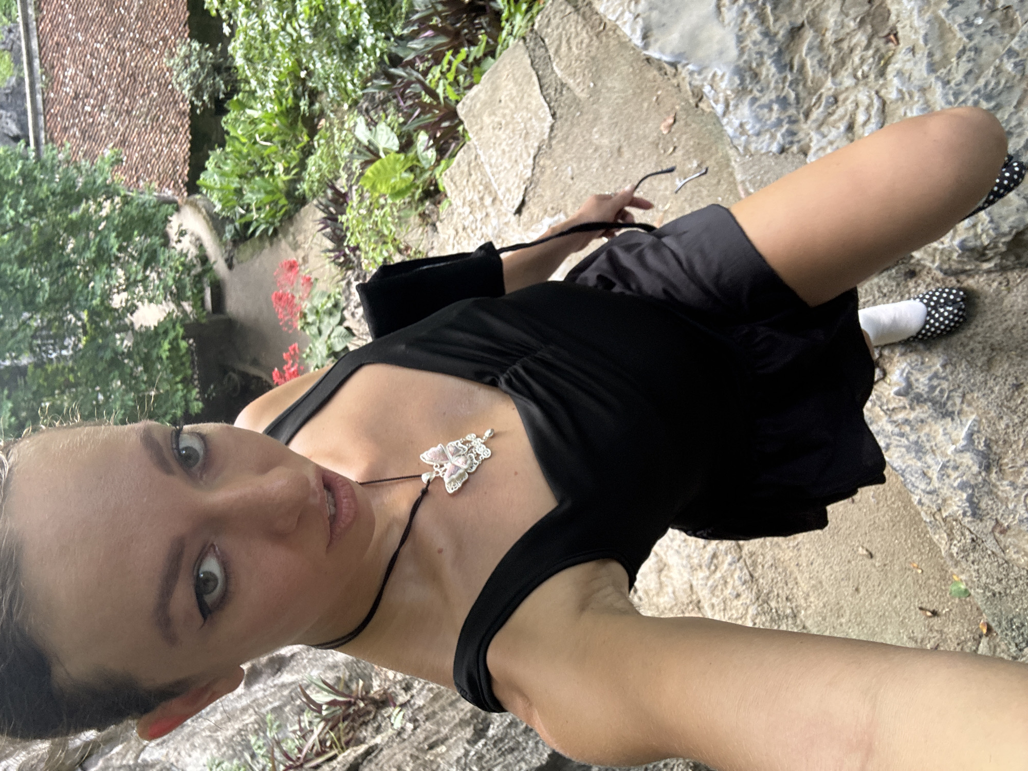 A high-angle, medium shot captures a woman in a black dress and polka-dot shoes on a stone staircase outdoors. Her hair is pulled back, and she wears a butterfly pendant. She holds a pair of glasses in her right hand. The background reveals lush greenery and a red-tiled roof. The scene appears to be in a garden setting, potentially within Nam Hoa Lư Ward, Vietnam, given the architectural style and vegetation.
