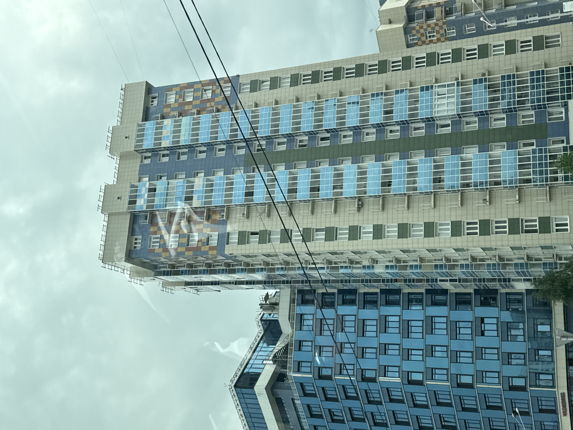 The image shows two modern apartment buildings in Krasnoyarsk, Russia, under a cloudy sky. The building on the left is primarily clad in blue glass, with sections of dark grey. It features multiple stories with repeating window patterns. The building on the right is taller and more colorful, with sections of light beige, blue, green, and a patterned mosaic of yellow and brown squares. Both buildings have balconies with white railings. Several power lines stretch across the upper portion of the image, obscuring the sky in places. The lower portion of the building on the left shows a storefront with the text "WILDBERRIES". There are no people visible in the image. The overall impression is of a contemporary urban landscape.