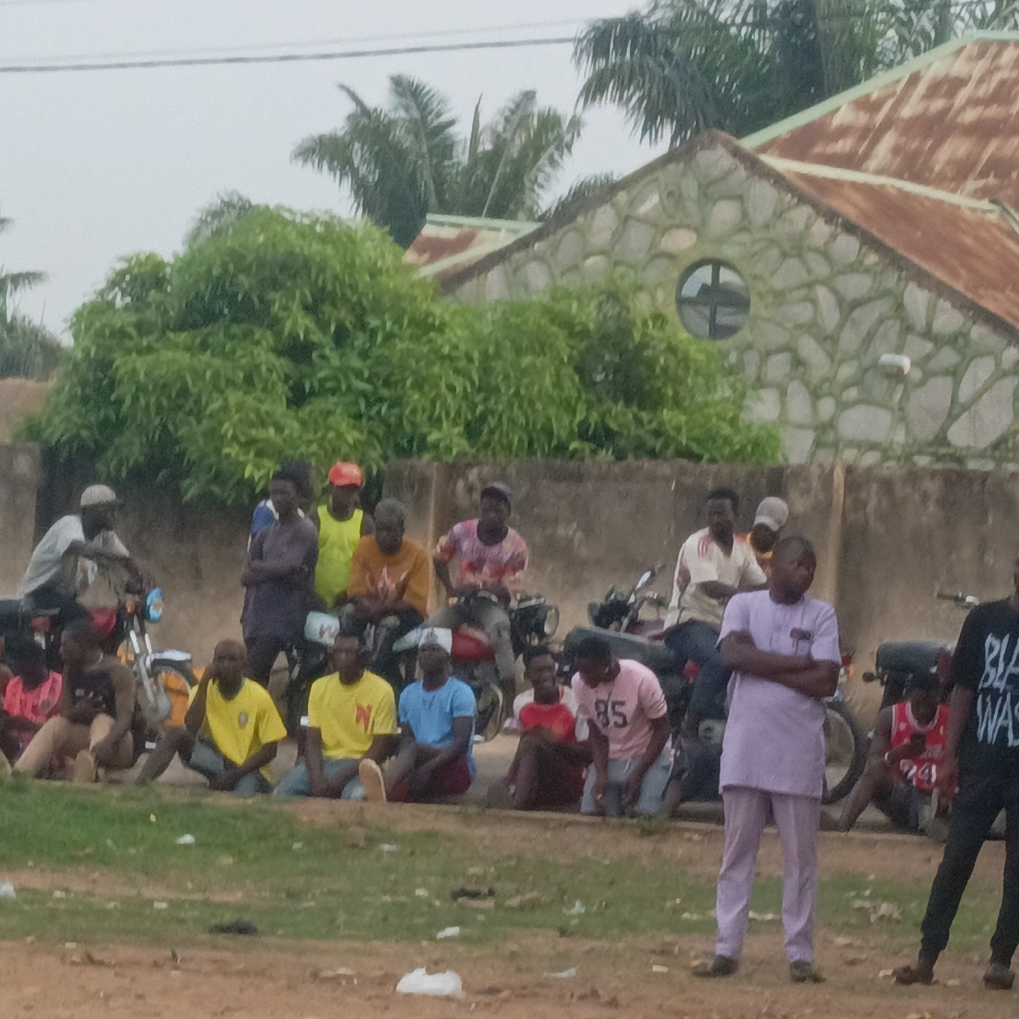 This image depicts a casual outdoor gathering of a group of men in Akwanga, Nigeria, under an overcast sky. The setting appears to be a semi-urban or rural area with a dirt ground interspersed with patches of grass.
Approximately 15-20 men are visible, mostly young to middle-aged adults. Many are sitting directly on the ground, some are seated on motorcycles, and a few individuals are standing. Their attire is casual, consisting mainly of t-shirts and trousers. Notable visible text includes a red "N" on a yellow t-shirt worn by a man sitting on the ground, the number "85" on a pink t-shirt worn by another man also sitting, and partial white text "BLA WAS" on a black t-shirt worn by a standing man on the far right.
Several motorcycles are parked or being used for seating, suggesting they are a common mode of transport or are used for work by some individuals. In the background, behind a low concrete wall, stands a building with a unique facade made of light-colored stones, featuring a distinctive circular window. The building is topped with a rusty, corrugated metal roof. Lush green trees and bushes, along with palm trees, frame the scene against the grey sky, indicating a vibrant natural environment. The overall impression is a relaxed social congregation or a waiting area.