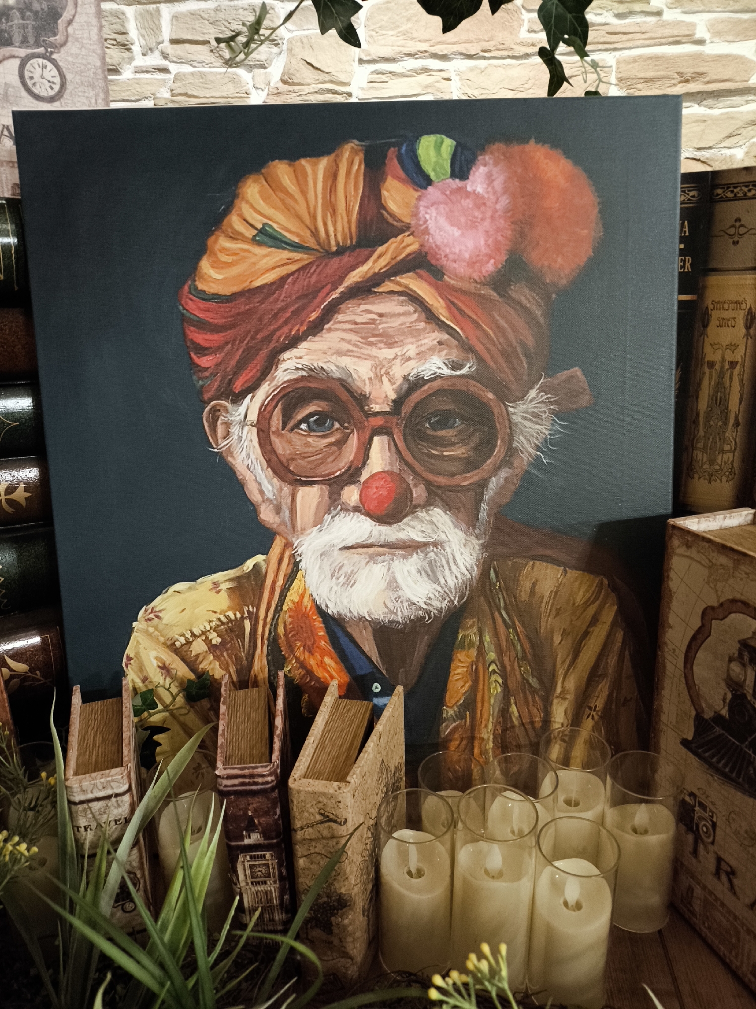 The image displays a painting of an elderly clown with a red nose and large, round glasses. He wears a colorful turban and a patterned jacket. The painting is positioned in front of a brick wall and is surrounded by decorative items including books, plants, and candles. The scene is lit with artificial lighting, casting soft shadows. No specific location in Moscow, Russia, is identifiable from the visual information provided.
