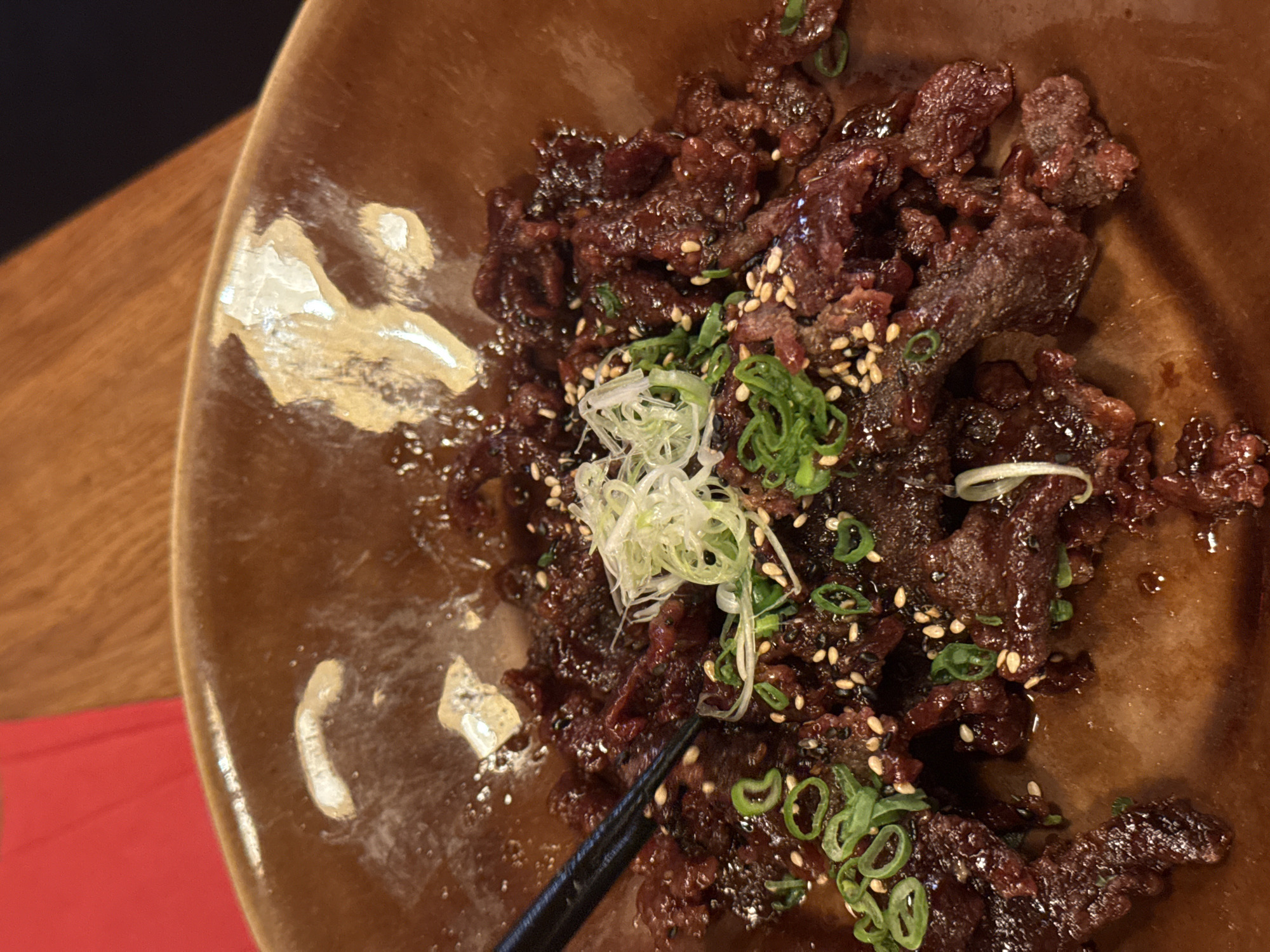 A close-up, overhead shot shows a brown ceramic bowl filled with pieces of dark, glazed meat, most likely beef, garnished with sliced green onions, white shredded scallions, and sesame seeds. A pair of black chopsticks rest in the lower left of the bowl. The bowl shows some light reflection and sits on a wood-grained surface, with a hint of a red object appearing near the bottom left corner. The city of Sofia, Bulgaria is not discernible from the image.