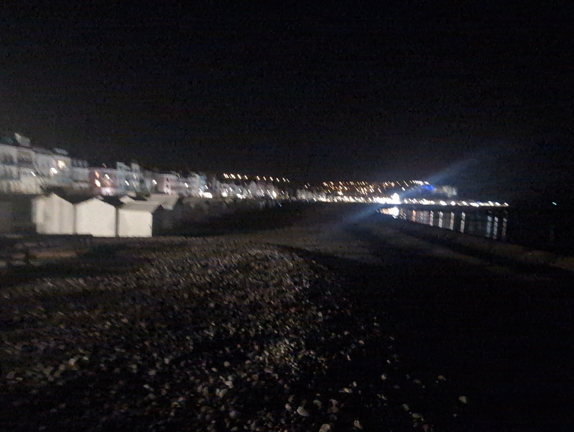 The image depicts a nighttime scene of the waterfront promenade in Mers-les-Bains, France. In the foreground, a dark, pebbled beach extends into the frame. To the left, three white beach huts are visible, appearing as rectangular structures against the darkness. Beyond the beach huts, a line of buildings with illuminated windows stretches along the promenade, forming a streetscape with intermittent lighting. The architecture suggests a row of houses or hotels typical of a seaside town. Further in the distance, the lights of the town continue along the coastline. On the right side of the image, the sea is present, reflecting the distant lights and exhibiting a slight shimmer. A pier or jetty structure is also visible extending into the water, with vertical elements illuminated by lights. A bright beam of light, possibly from a searchlight or powerful spotlight, cuts through the night sky from the right side of the frame, illuminating a section of the buildings. The overall impression is one of a quiet, illuminated coastal town at night.