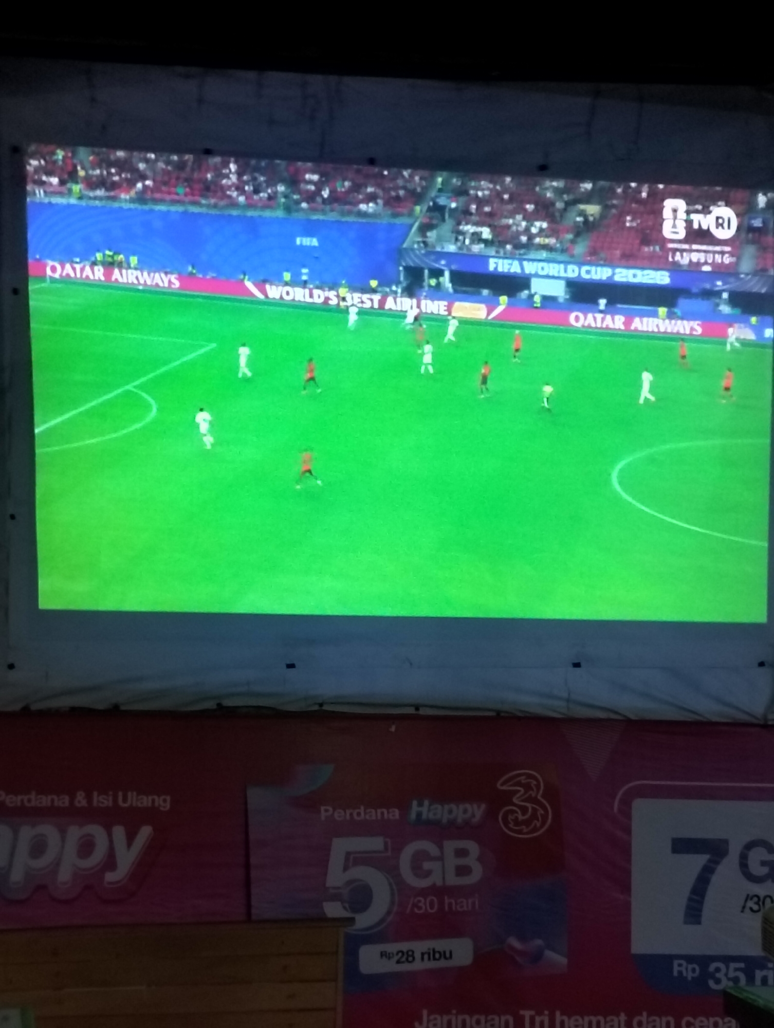 The image shows a projection screen displaying a soccer match, likely part of the FIFA World Cup 2026, with "QATAR AIRWAYS" and "WORLD'S BEST AIRLINE" visible as advertisements on the field. The screen is set up outdoors, possibly in the city of Lhokseumawe, Aceh, Indonesia, given the context of the advertisement below for "Perdana & Isi Ulang Happy 3". The bottom of the image displays promotional material for "Happy 3" mobile services, featuring offers of "5 GB /30 hari Rp 28 ribu" and "7 GB /30 hari Rp 35 ribu". The overall scene suggests an outdoor public viewing or event related to the soccer tournament and a telecommunications promotion.