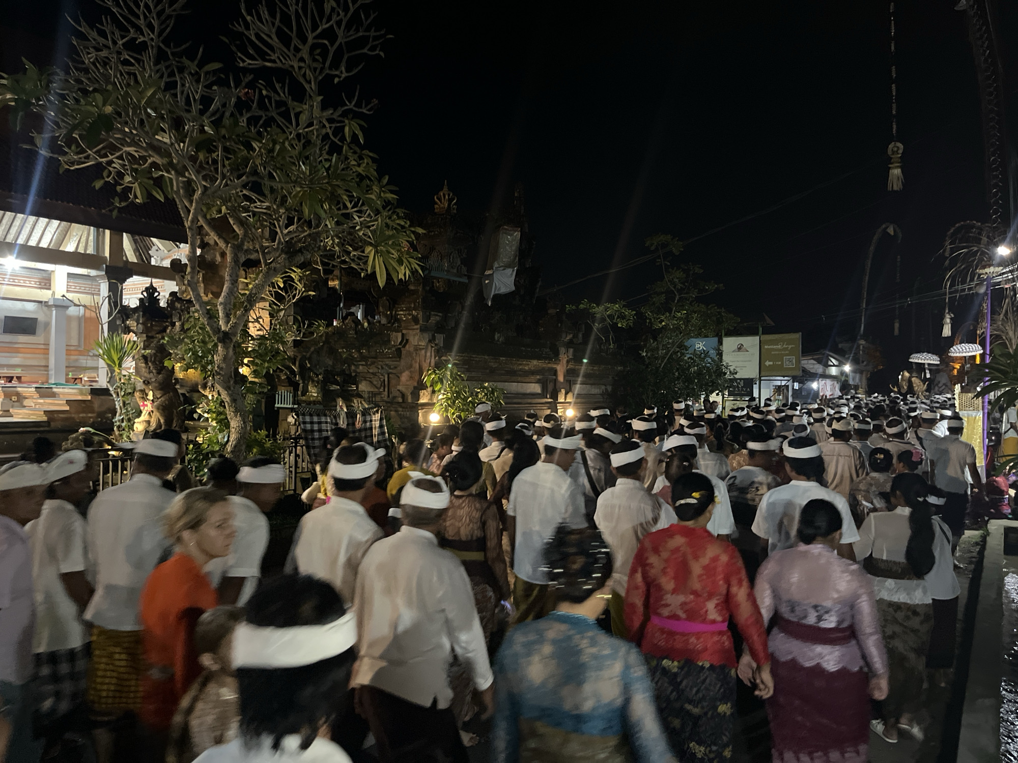 The image depicts a nighttime procession of people in what appears to be a village or temple setting. The subjects are a large group of individuals, many wearing white shirts and headbands, suggesting a formal or religious occasion. Some women are wearing traditional attire, including sarongs and intricately patterned tops. The scene is illuminated by artificial lights, casting shadows and creating a dramatic atmosphere.
The procession is moving along a path or street, with buildings and trees visible on either side. The architecture suggests a Southeast Asian location, and the presence of ornate doorways and traditional decorations reinforces this. Hanging decorations, possibly religious or ceremonial, are visible in the background. The overall mood is one of solemnity and community participation.
Notable details include the time of day, which is clearly night, and the presence of a barren tree with sparse foliage in the foreground. The weather appears to be clear, as there are no signs of rain or strong winds. Location cues include the architectural style and the attire of the people, which are consistent with cultural practices in Indonesia, specifically the Ubud District.
There are no discernible emotions expressed clearly on the faces of the individuals due to the distance and lighting conditions. The activity appears to be a procession or parade, possibly part of a festival or religious ceremony. The image captures a moment of collective movement and cultural expression under the night sky. No visible text is clear enough to be read.