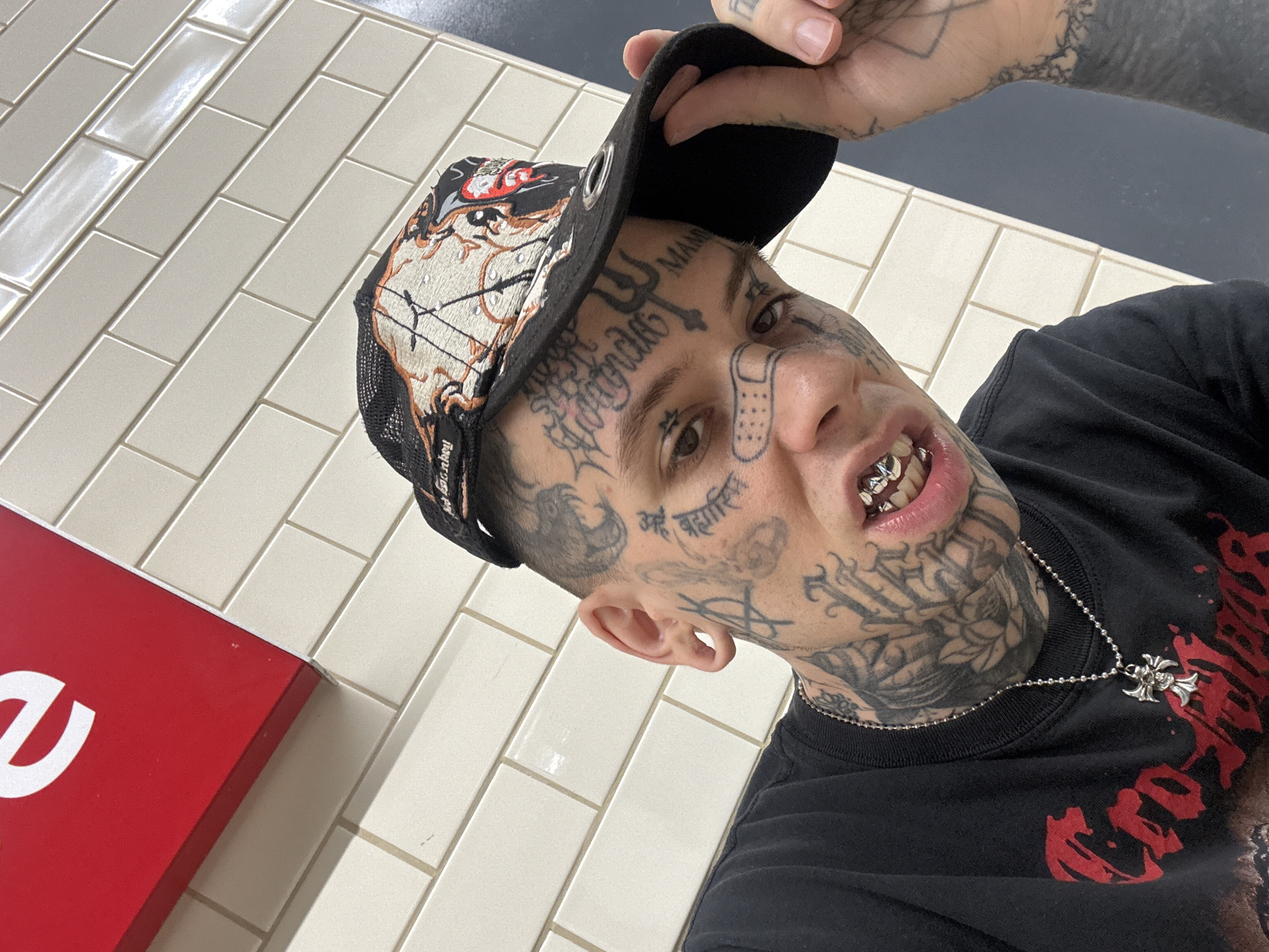 A close-up, slightly angled, indoor selfie of a heavily tattooed man with facial tattoos, wearing a black baseball cap with a design on the front, a black t-shirt, and a silver necklace with a cross pendant. He has silver grills on his teeth and is touching the brim of his cap with his right hand. The background is a wall of rectangular white tiles. A red rectangular object is partially visible in the lower left corner. The location cannot be determined based on the visual information provided.