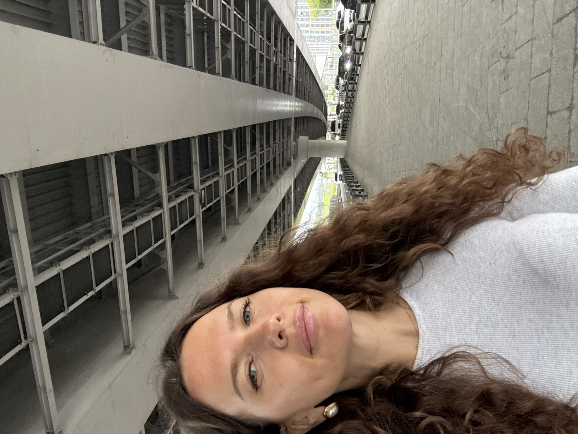A woman with long, wavy brown hair and blue eyes is taking a selfie under a large concrete overpass. She is wearing a light gray ribbed long-sleeve shirt and a gold earring with a pearl. The overpass has a complex network of beams and supports above. To the right of the woman, a paved walkway runs alongside a road where several cars are visible driving in both directions. In the background, tall buildings and some trees can be seen. The scene appears to be in an urban environment, possibly Moscow, Russia, given the architecture and the presence of a multi-level road structure.