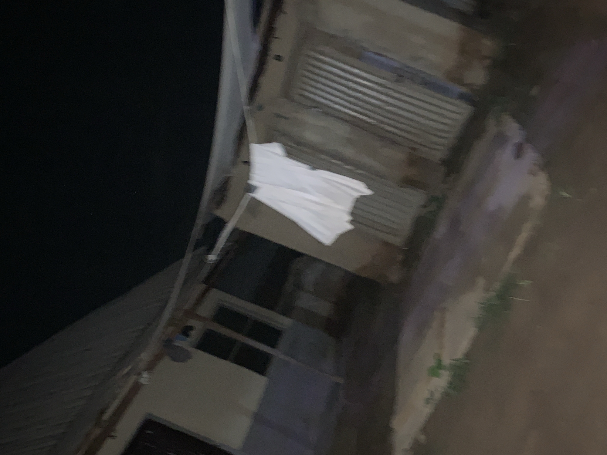 The image, taken at night, shows a view of a building exterior. The location is presumed to be Keffi, Nigeria, based on the prompt. A white cloth or piece of clothing is draped over a structure resembling a clothesline or railing. The building has a corrugated metal section, potentially a roof or wall. There is a window visible. The environment appears to be dimly lit, possibly a street or yard adjacent to the building. No people are visible.