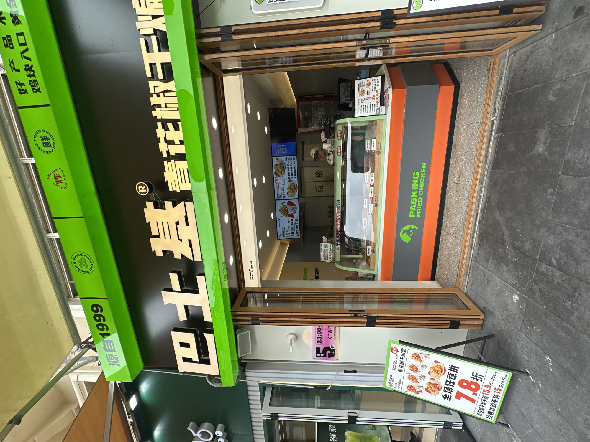 Here's a description of the image:
The image shows the front of a "PASKING FRIED CHICKEN" restaurant. The storefront is in a city environment, indicated by the visible pavement.
The restaurant has a green and black sign with Chinese characters and the "PASKING FRIED CHICKEN" logo. Below the main sign is the founding date 1999. The entrance features folding glass doors, through which the interior and staff members can be seen.
A sandwich board displays a promotion of 7.8, offering discounts on items like the Qinghua Pepper and dry-fried series. Another sign indicates closing time as 23:00 (11 PM).
The location is likely in China. "PASKING FRIED CHICKEN" is a brand that originated in China, and the menu visible inside the store has menu items like Qinghua Pepper and dry-fried series as well.