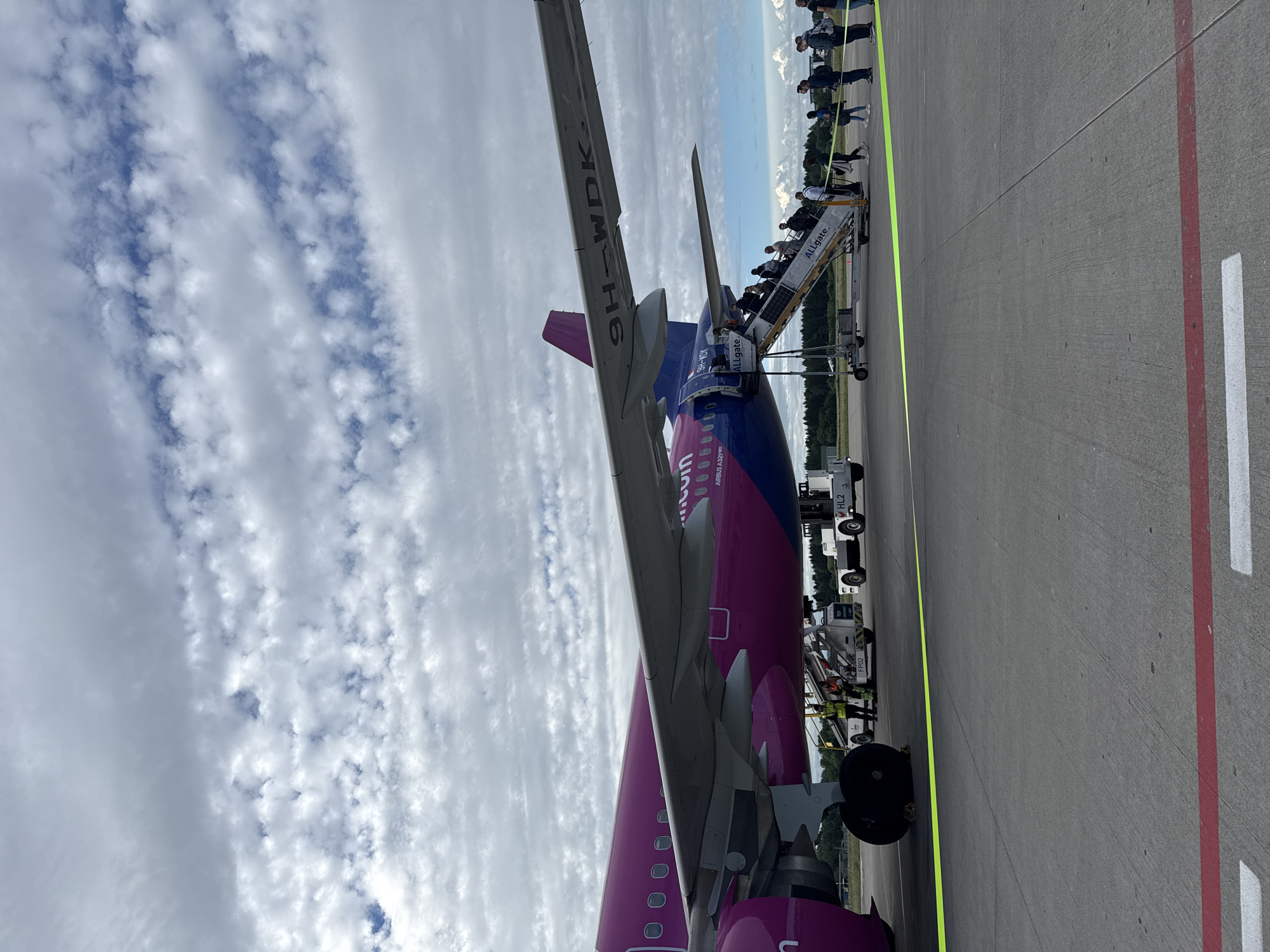 An Airbus A320-200 aircraft with registration 9H-WDK, operated by Wizz Air, is parked on the tarmac at Memmingen Airport (FMM) in Künersberg, Germany. The aircraft is predominantly pink and blue, with the Wizz Air logo visible. Passengers are boarding the aircraft via mobile stairs, indicated by the "ALLGATE" branding on the stairs. Several individuals, including passengers and ground crew, are present near the plane. The scene takes place under a cloudy sky with patches of blue. The ground is paved asphalt, marked with yellow and red lines. Trees and distant buildings can be seen in the background.