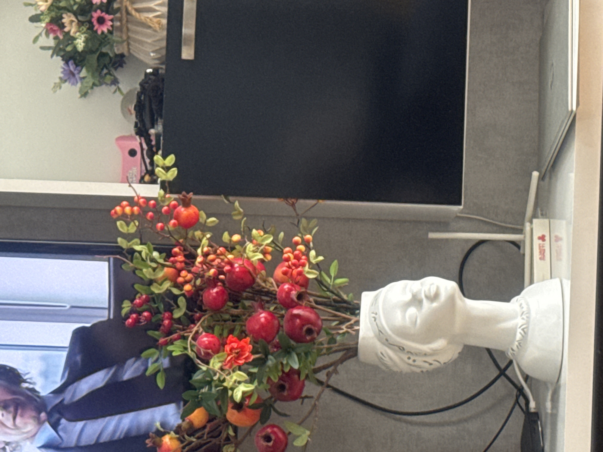 The image displays a well-lit indoor scene, likely a contemporary living or office space, showcasing a mix of decorative elements and electronic devices. The photograph is oriented vertically, appearing as if shot in portrait mode and then rotated, or the scene itself is arranged vertically.
In the foreground, a striking white ceramic bust, sculpted in the form of a woman's head with intricate detailing on her hair and base, functions as a unique vase. From the head of this bust, a lavish artificial arrangement of vibrant red pomegranates, clusters of smaller red and orange berries, and scattered orange flowers with green foliage and brown branches overflows, creating a dynamic visual centerpiece.
Behind this floral display, a large, rectangular black screen, likely a television or computer monitor, dominates the background to the left. A partial reflection or image on the lower-left corner of this screen shows a person, possibly a man, wearing a dark suit jacket over a striped shirt, with only a side profile of his face visible.
To the right of the ceramic bust, on a white shelf, a small white rectangular device, consistent with a Wi-Fi router, is positioned with various black electrical cables snaking around it. Adjacent to this device, a few white, vertically stacked objects, possibly books or boxes, are visible; one spine shows indistinct red text.
In the upper-left portion of the image, another decorative item, an artificial floral wreath or hanging basket with pink, purple, and white flowers and green leaves, is mounted against a light wall. Below this, a small pink object with a handle or nozzle is partially discernible on a shelf.
The scene is composed of various objects on a textured light grey surface and white shelves against a matching light grey wall, suggesting a thoughtfully arranged interior. No specific time of day or weather conditions are discernible, and there are no direct visual cues within the image to indicate its location as Tbilisi, Georgia.