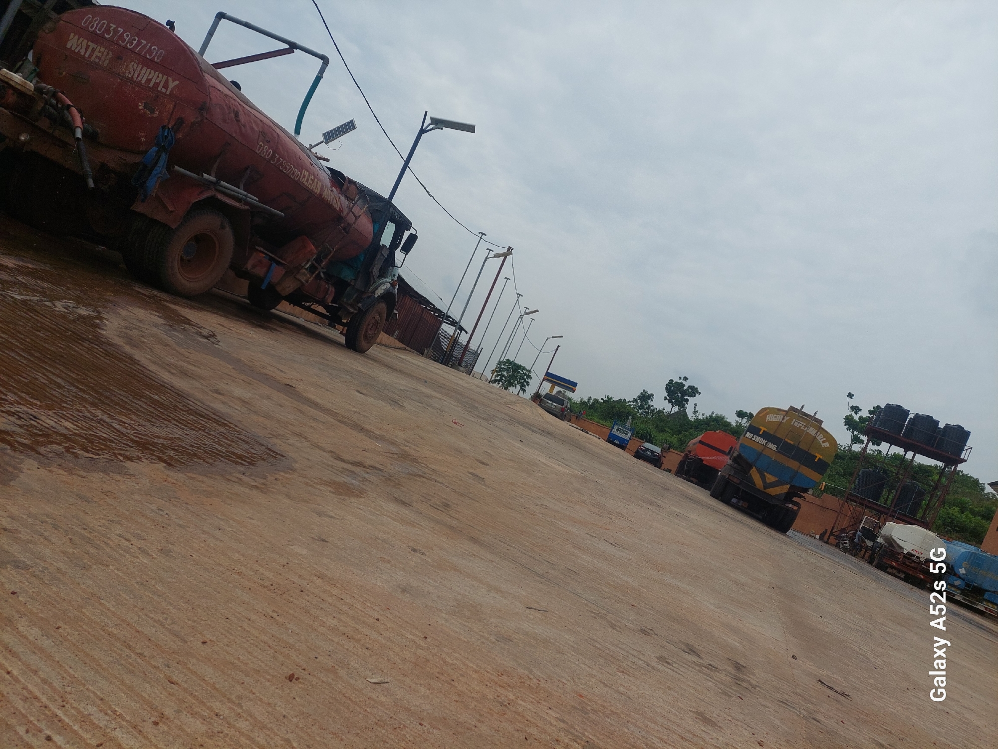 The image depicts a street scene in Awkuzu, Nigeria. The foreground is dominated by a textured, sandy-brown surface, possibly a road or pathway, with diagonal striations suggesting it has been raked or weathered. A portion of this surface appears wet and reflective.
In the mid-ground, several water tanker trucks are parked in a line. The closest and most prominent is a large, red tanker with "WATER SUPPLY" printed in white lettering and a phone number "08033793150". Another tanker behind it is blue and yellow with stripes and visible text that includes "NO SMOKING" and "HIGHLY INFLAMMABLE". Further down the line, other tankers in various colors can be seen, some with additional markings. The trucks are positioned at an angle, suggesting the photo was taken from a slightly elevated viewpoint or that the trucks are parked on a slope.
The background features a line of trees and vegetation, hinting at the presence of a green area or a more rural setting within the city. The sky above is overcast and a pale blue-grey. The overall impression is one of utility vehicles parked on a street, likely for a water distribution service. The image is oriented vertically, with the bottom of the frame appearing on the left. A watermark in the lower left corner reads "Galaxy A52s 5G".
