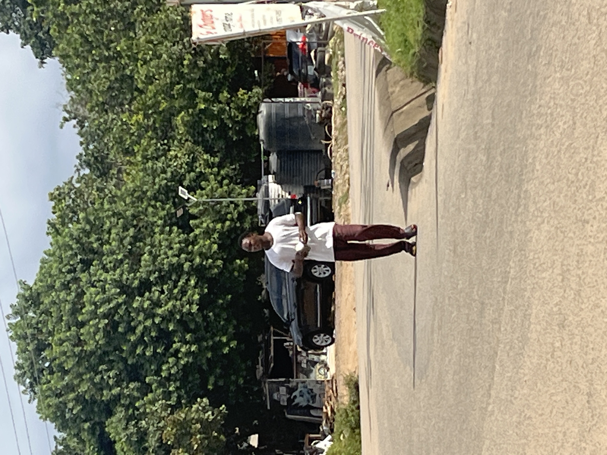 A young Black man, wearing a white t-shirt and maroon pants, walks across a paved road in Abuja, Nigeria. He is holding a white object in his right hand. In the background, a dark-colored SUV is parked on the side of the road, next to several large black water tanks. A small structure, possibly a vendor's stall, is located behind the SUV. Dense green trees form a backdrop to the scene. A white signpost with red lettering stands to the right of the image, partially obscured by a grassy embankment. The sky is overcast, with some power lines visible in the upper portion of the frame. The road appears to be a two-lane street, with a concrete median in the foreground.